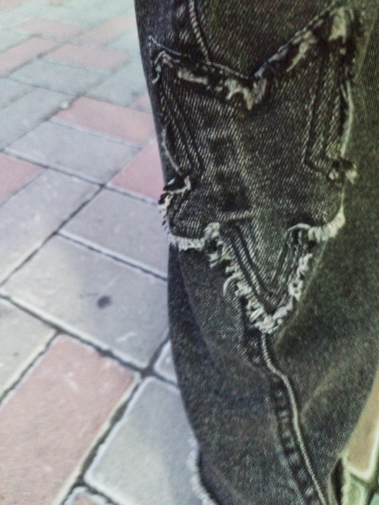 The image displays a close-up view of distressed denim fabric, likely from a pair of jeans, featuring a star-shaped patch with frayed edges and visible stitching. The texture of the denim is rough and appears worn. In the background, a paved surface composed of rectangular stones in varying shades of gray and pink is visible, suggesting an outdoor urban environment. The lighting is even, indicating daytime conditions. No specific landmarks or elements definitively place the scene in Surgut, Russia.