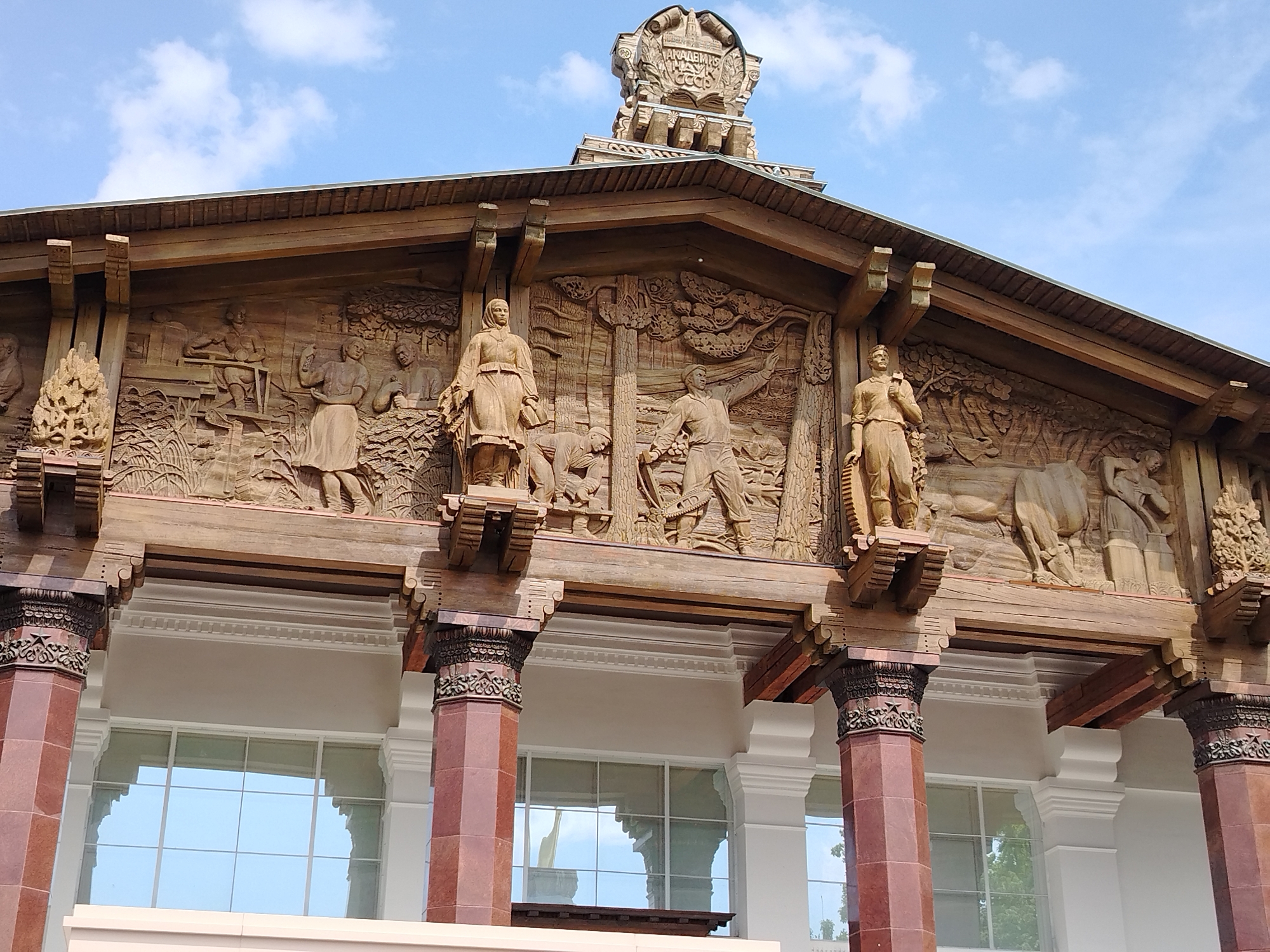 The image showcases a portion of the facade of a building with a prominent architectural style, featuring intricately carved wooden panels depicting scenes of agricultural and industrial activities. The building is adorned with decorative elements, including statues of figures holding tools or produce, and elaborate relief carvings of people engaged in farming, logging, and other productive endeavors. Below the wooden panels, the building has large windows and ornate columns in a reddish-brown hue with carved details at the top. Above the paneling is a tower-like structure with similar carvings. The overall aesthetic suggests a structure built during the Soviet era, possibly as part of a larger complex such as an agricultural exposition or a public building. The setting appears to be outside, with a clear sky providing natural light.