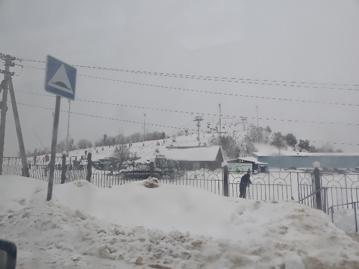 This image depicts a snow-covered winter landscape in Курово, Russia, under an overcast sky. In the immediate foreground, large, dirty snow piles occupy the lower portion of the frame, with a vehicle interior component visible in the bottom left corner. A blue square road sign, indicating a speed bump with a white triangle and black icon, stands on a pole in the mid-left. To its left, a utility pole supports several parallel power lines traversing the upper image.
A dark, ornamental metal fence, partially buried in snow, stretches horizontally across the midground. Behind this fence, a person wearing a dark blue jacket and pants is observed next to a snowdrift, appearing to shovel or interact with the snow. Beyond the fence, several buildings are visible, including a rustic wooden structure with a dark roof on the left and a larger, lighter-colored building with a blue section to the right. A parking area containing multiple vehicles is present behind these structures.
The background is dominated by a snow-covered ski slope featuring multiple ski lifts and their supporting towers. Numerous individuals, identified as skiers or snowboarders, are dispersed across the slope, actively engaged in winter sports. Scattered bare trees are visible on the snow-laden hillside.