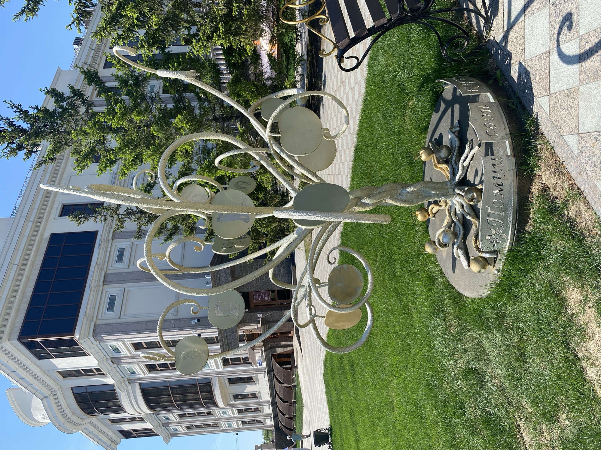 A full shot depicts an outdoor installation in Tyumen, Russia, against the backdrop of a modern building and lush green grass. The installation, in the foreground, is a stylized metal tree with circular discs resembling leaves, some of which bear images. The tree is painted in a light color, possibly white or cream. At the base of the tree, there is a circular stone or concrete platform with inscriptions in a language that appears to be Russian. There are metal decorative elements, including what appears to be a representation of apples, at the base of the tree. Adjacent to the installation is a paved walkway constructed of light-colored stones, with a bench visible in the background. The modern building behind the installation has a facade of alternating bands of light-colored stone and dark glass windows. Green trees are partially visible between the installation and the building, adding to the scene's verdant feel. The sky is blue with clear weather.