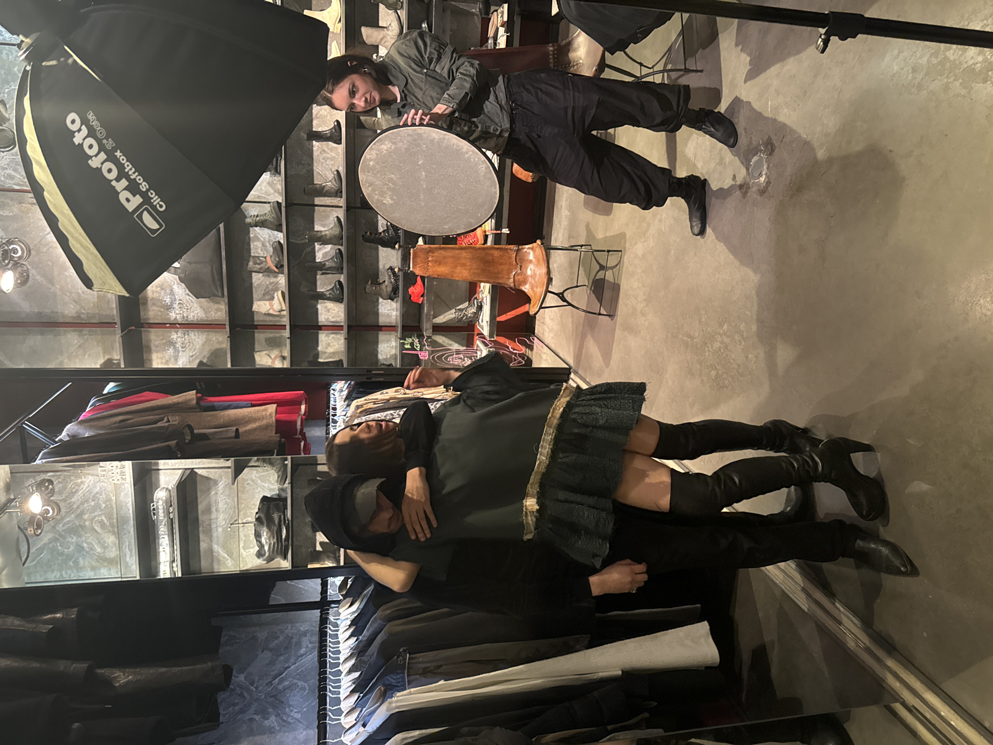 The image depicts a fashion shoot in progress. A woman in work clothes and a hair tie stands with a large reflector while a softbox light sits overhead on a stand. A second woman is being held by a man, both wearing clothes being displayed in the surrounding racks. The location appears to be a clothing store featuring displayed racks of clothing and accessories. Based on the style of clothing and the overall ambiance, the location is likely in Paris, France.