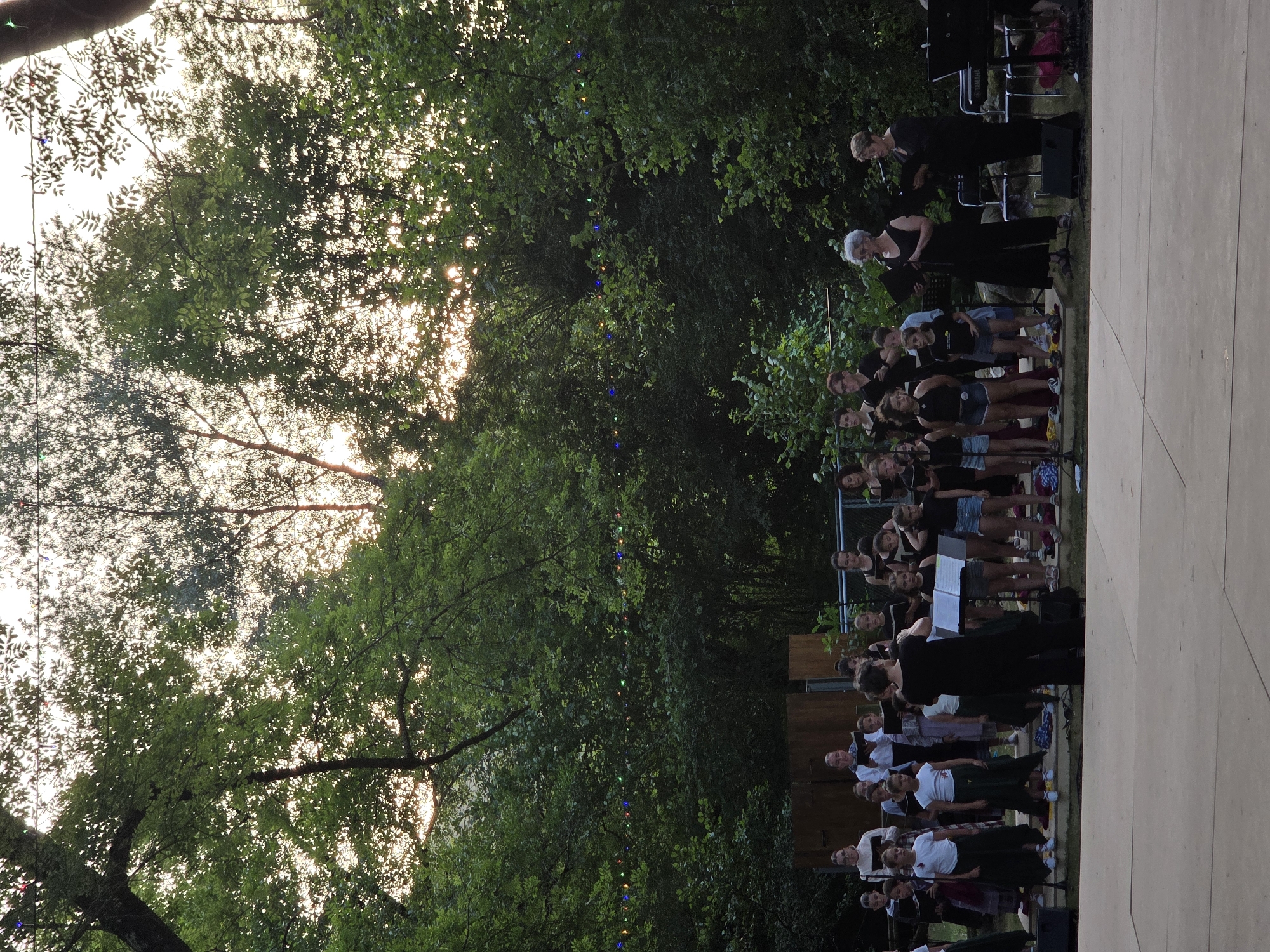 The image depicts an outdoor choir performance taking place in a park-like setting. A group of children and adults, dressed in performance attire, stand on a stage, holding sheet music. Some are singing, while others appear to be conducting or observing. The background is filled with lush green trees, and string lights are visible overhead, suggesting an evening or twilight performance. The stage itself is a flat, light-colored surface. The overall atmosphere appears to be communal and focused on the performance.