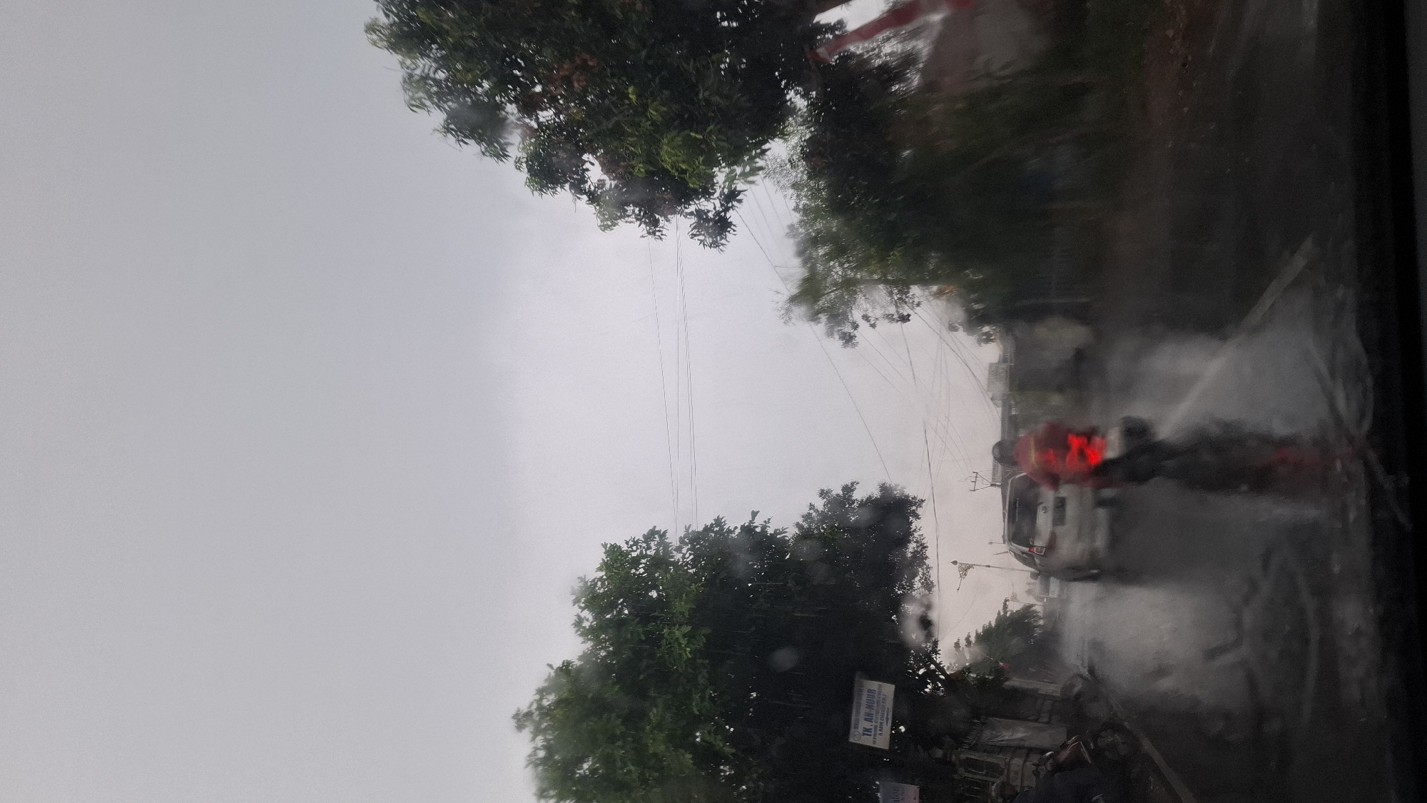 The image depicts a street scene in Cilaku, Indonesia, during a heavy rainfall. The perspective is from inside a vehicle, looking forward through a wet windshield. The road is wet and reflective, with puddles of water visible. In the foreground, a motorcycle rider wearing a red jacket is visible, kicking up water as they move away from the camera. To the left, a white car is also traveling on the road. The environment is characterized by lush green trees on either side of the street. There are also buildings and power lines present. The sky is overcast and gray, indicating poor weather conditions. The overall impression is of a typical Indonesian street scene during monsoon season.