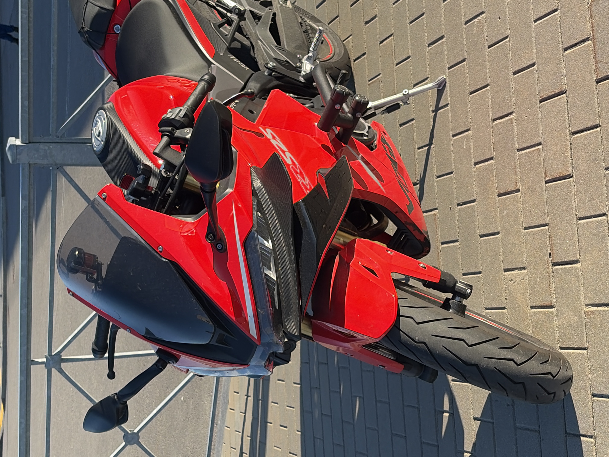 A red sport motorcycle is parked on a paved surface in Saint Petersburg, Russia. The motorcycle is positioned in the foreground, facing the viewer. It is a sunny day, casting shadows on the ground. The background features a metal fence and a road. The motorcycle has a sleek design with black accents and carbon fiber detailing. The front wheel, handlebars, mirrors, and a portion of the fairing are clearly visible. The motorcycle appears to be stationary and unattended. The paved surface consists of rectangular brick-like stones laid in a pattern.