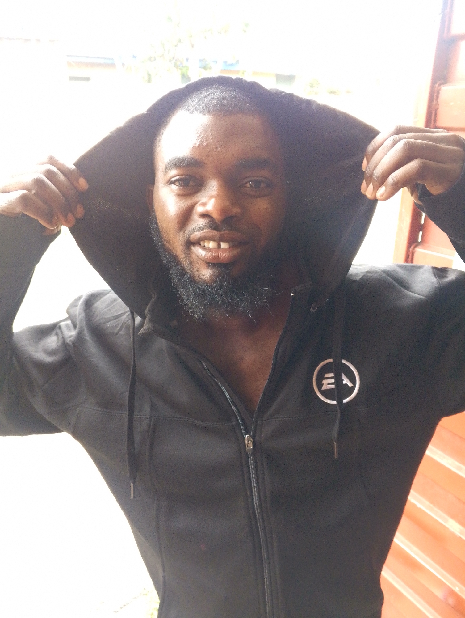 A dark-skinned man with a beard is posing in a black hooded sweatshirt. The hood is pulled up and held open by his hands on either side of his head. The sweatshirt has a circular logo on the left breast with the letters "EA" inside. The man is smiling, revealing a slight gap between his upper front teeth. The background is brightly lit and indistinct, but suggests an outdoor setting possibly in the city of Jos, Nigeria. The orange wall appears to be a fence.