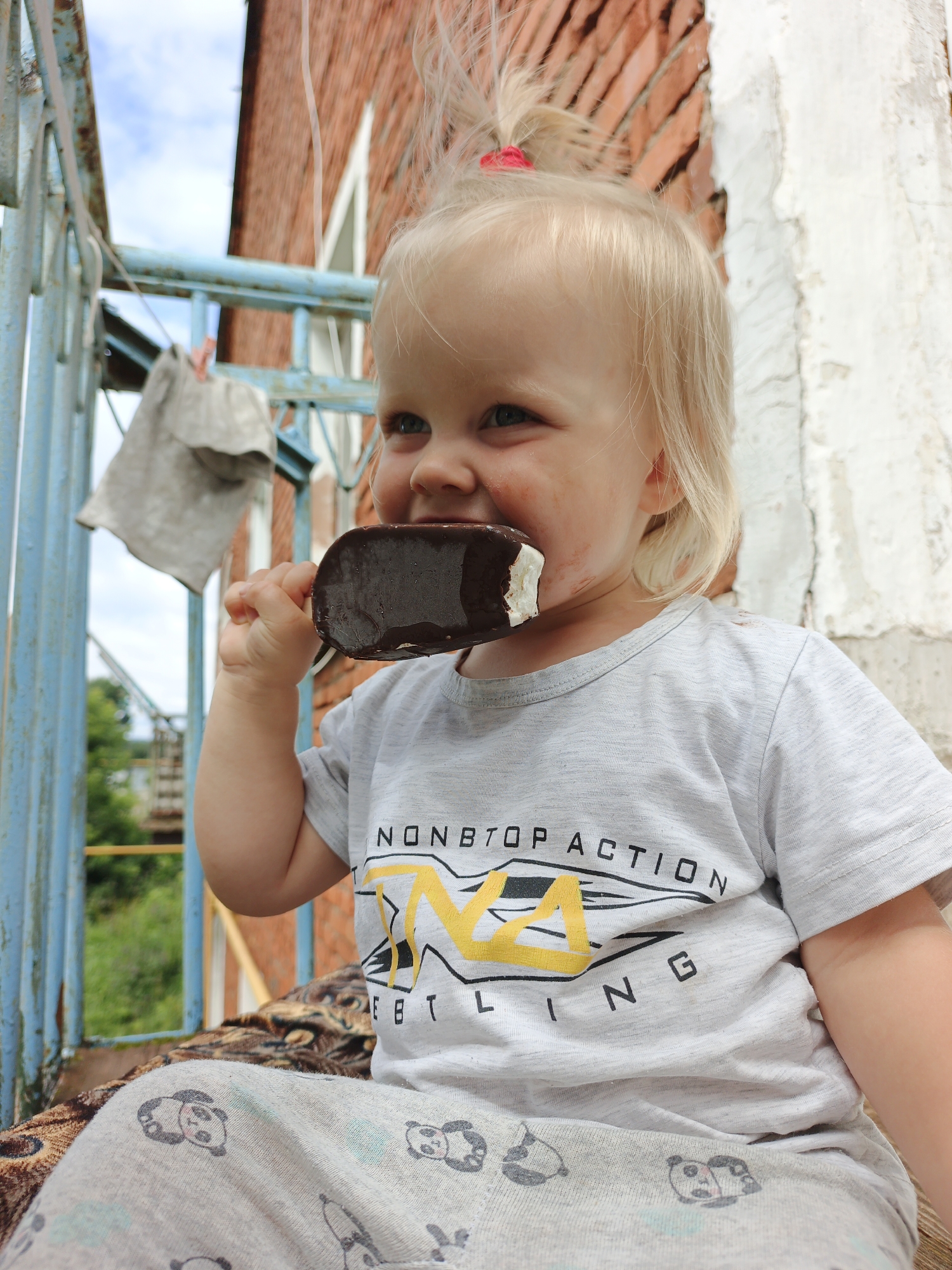 A blonde-haired toddler with blue eyes is seated outdoors, holding a chocolate-covered ice cream bar to their mouth with their right hand. The child is wearing a grey t-shirt with a graphic design and panda-patterned sweatpants. Behind the child, a weathered blue railing is visible, along with a brick wall and a white-painted surface. The background suggests an outdoor setting, possibly a balcony or porch, with glimpses of greenery and a distant structure.