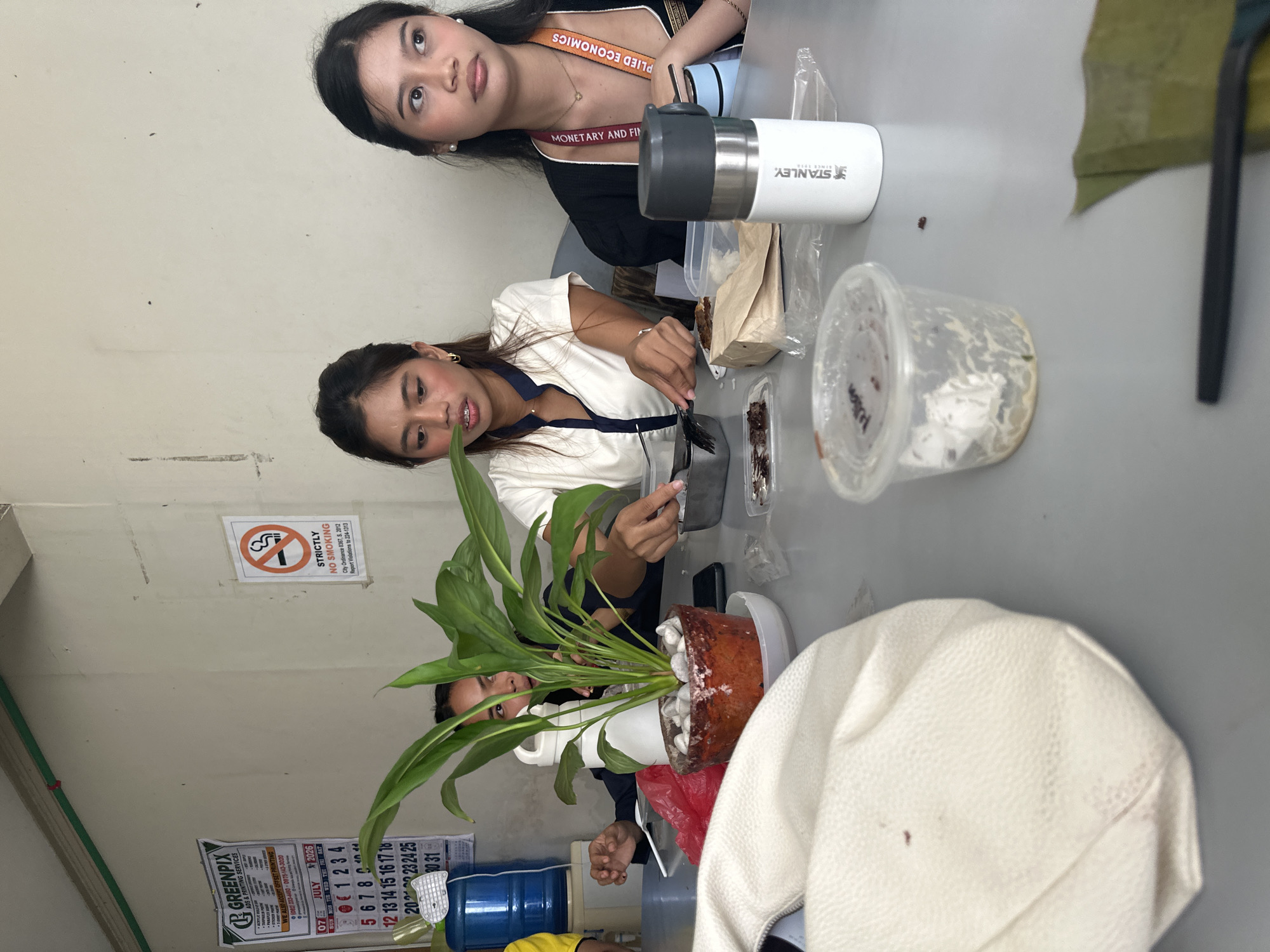 Three young women are seated at a gray table in what appears to be a modest establishment. On the wall behind them, a "Strictly No Smoking" sign is visible. A calendar from July 2026 is also displayed on the wall. The table is set with various items, including a potted plant, a water dispenser, a Stanley thermos, plastic containers, and food. The woman in the center is holding a fork and knife, seemingly engaged in eating or preparing food. The woman on the right wears a lanyard with text that appears to include "MONETARY AND FIN" and "PLIED ECONOMICS". The overall scene suggests a casual meal or break.