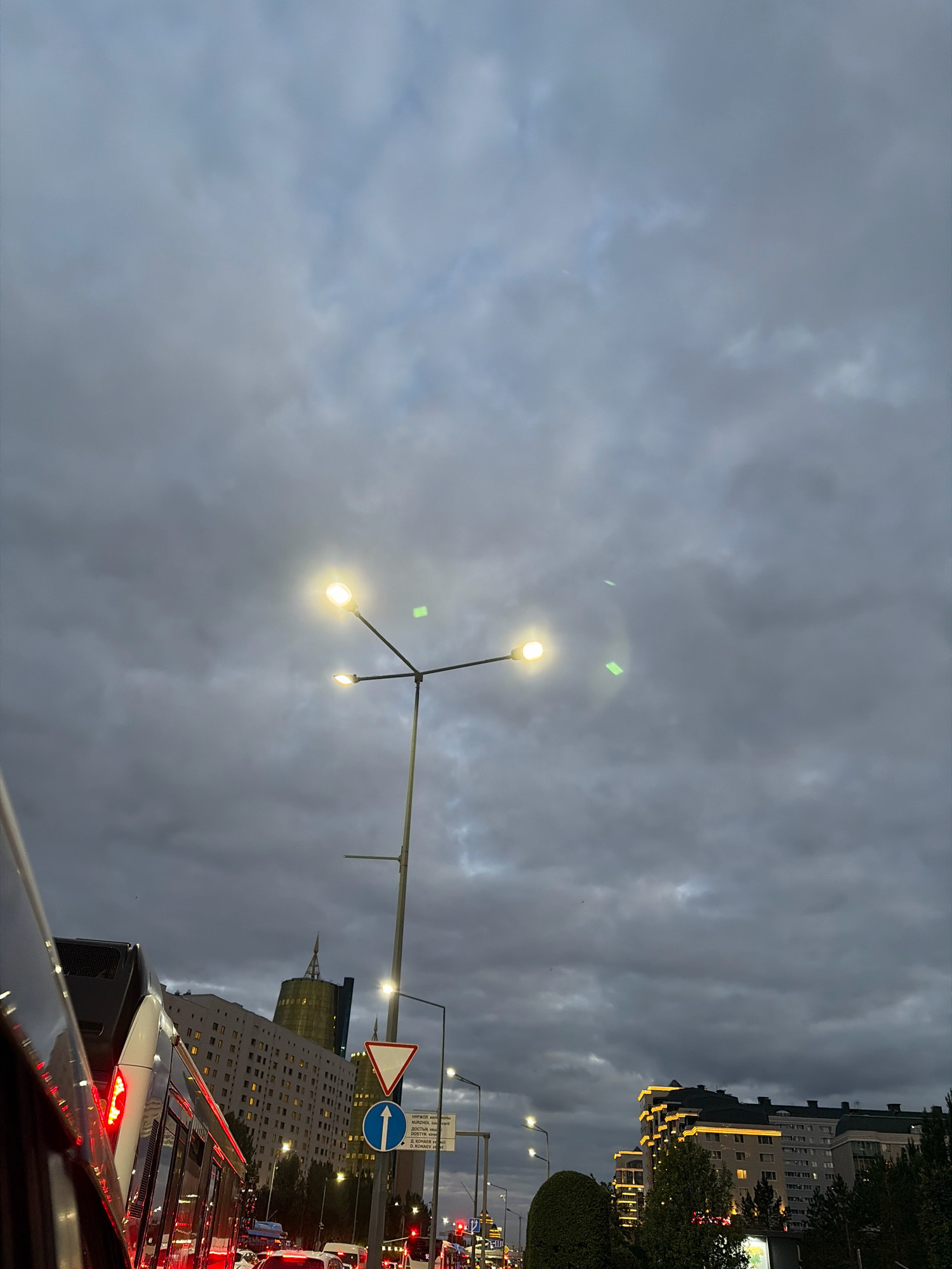 The image depicts a street scene in Astana, Kazakhstan, under a cloudy sky. A street lamp with two illuminated bulbs stands prominently in the foreground, casting a bright light against the dark clouds. Below the lamp, traffic signs are visible, including a yield sign and a directional sign. The scene is dominated by vehicular traffic, with the reflection of red brake lights visible on the side of a bus or tram in the left foreground. In the background, several buildings are visible, including a tall, modern building with a distinctive pointed roof and other apartment-style buildings. Some of the buildings are illuminated from within and along their upper edges, suggesting it is dusk or evening. The overall impression is of a busy urban environment at the end of the day, with overcast weather.