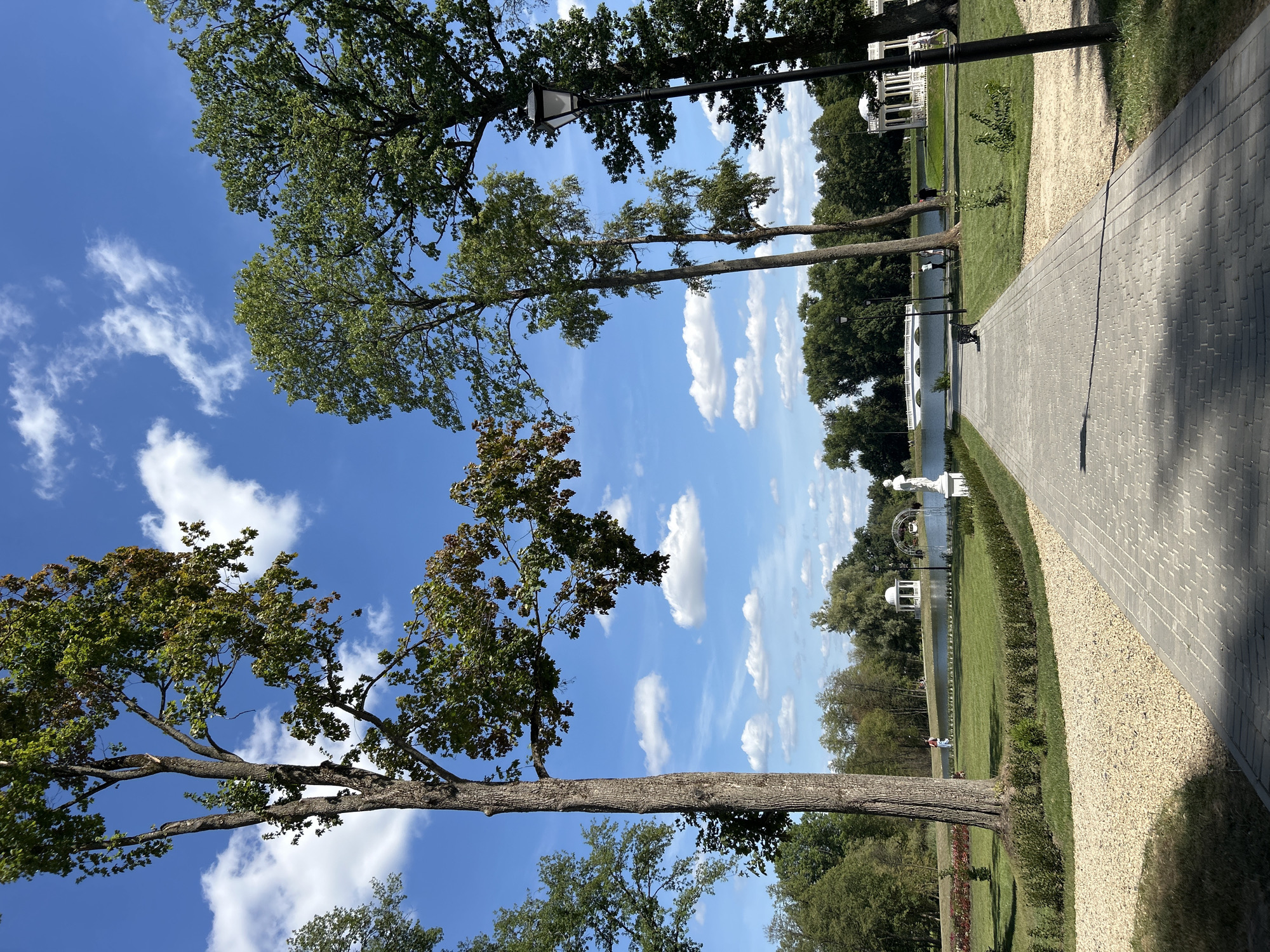 The image shows a paved walkway leading through a park towards a body of water. Several tall trees with green foliage line the walkway and surround the park. In the background, a white gazebo and a white arched bridge are visible across the water. The sky is blue with scattered white clouds. There are a few people scattered throughout the park, some walking and some sitting on benches. A statue stands to the right of the walkway near the water. The overall scene depicts a serene park environment. The location is identified as Dobasna Village Council, Belarus.