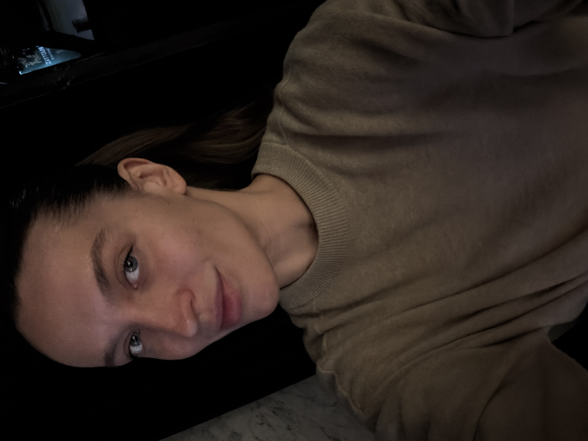 A young woman is pictured indoors, illuminated by dim lighting. She has fair skin, light eyes, and her dark hair is pulled back. She is wearing a beige crewneck sweater. The background is dark, with a glimpse of a screen displaying text in the top left corner. A marble or stone surface is visible in the lower left corner. There is no information about the city of Budva, Montenegro, to be seen in this picture.