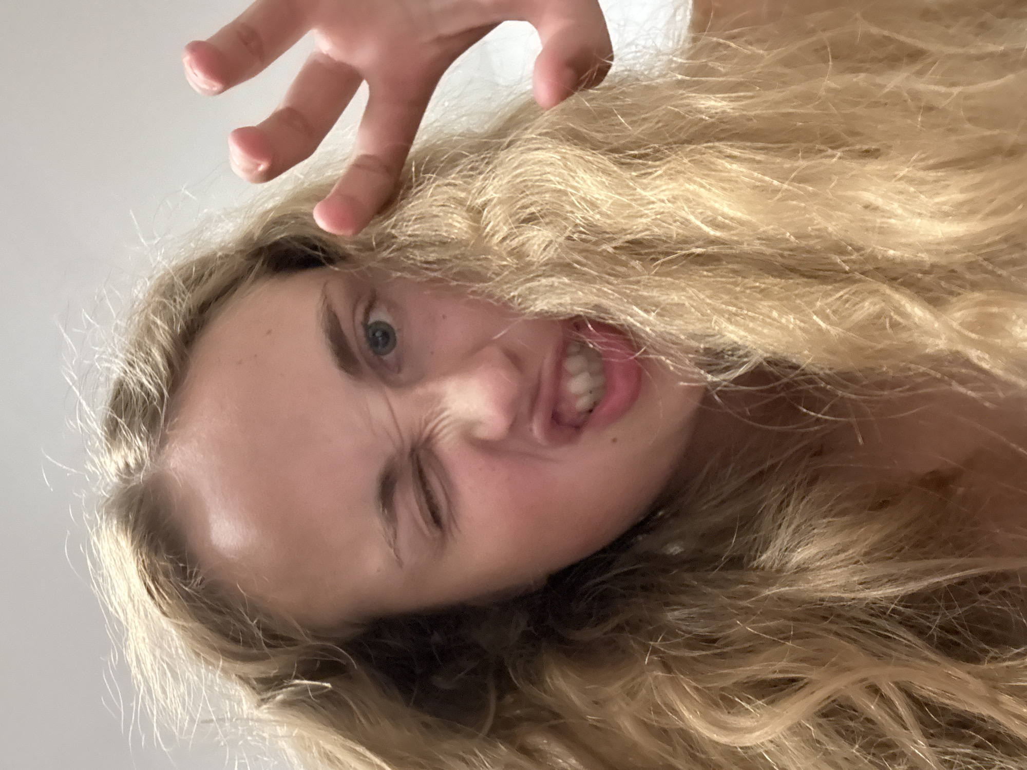 A young blonde woman is captured in a close-up, low-angle selfie. She is winking with her right eye and has her mouth open, revealing her teeth in a playful or mocking expression. Her left eye is open, showing a blue iris. Her long, wavy blonde hair is spread around her face. A hand with outstretched fingers is positioned in the upper right corner of the frame, partially obscuring her hair and face. The background is a plain, light-colored surface, possibly a wall or ceiling. The image appears to be taken indoors, with the lighting suggesting it is daytime. There are no discernible landmarks or environmental details that would place this image in Pula, Croatia.