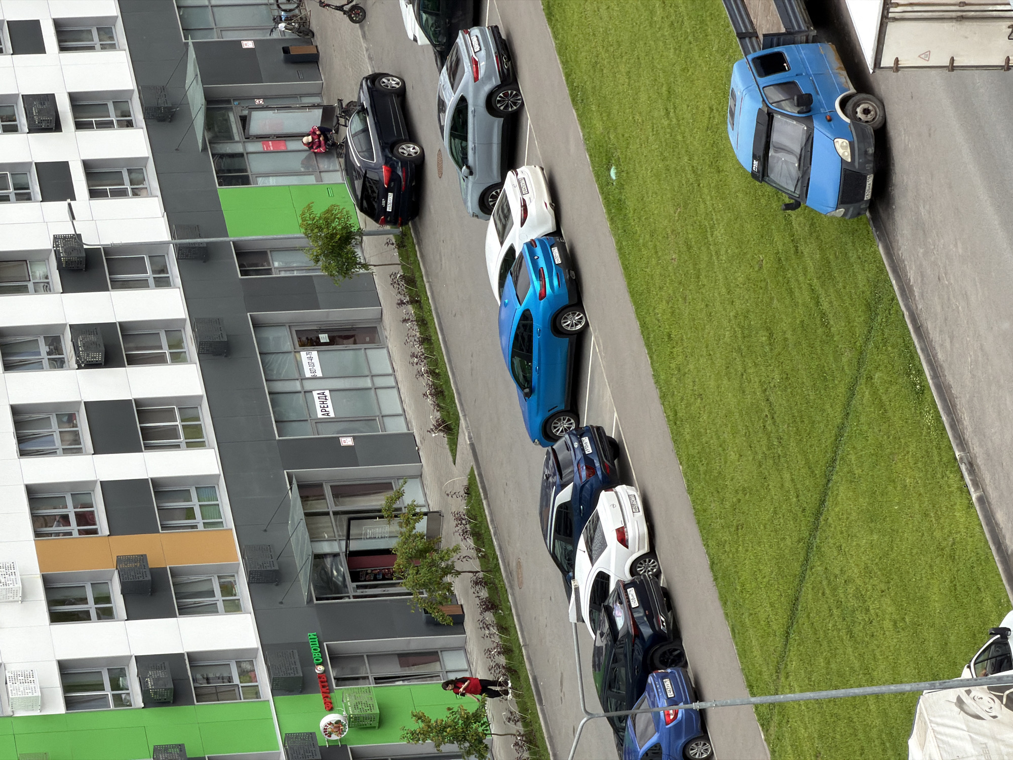 This is a high-angle, eye-level shot of a modern residential building with cars parked outside. The building is a multi-story structure with a facade featuring a combination of white, dark gray, and bright green panels, along with numerous windows. In front of the building, a paved area serves as a parking lot with several cars lined up. The cars are of various colors, including black, gray, white, and blue. A strip of green grass runs alongside the parking area.
In the foreground, on the right side of the image, a blue van is parked facing the viewer. A portion of a white truck or trailer is visible behind it. On the left side of the image, a woman in a red jacket and dark pants is visible, appearing to be engaged in some activity near a tree and a shopping cart.
Some visible text includes "АРЕНДА" (rent) on a sign on the building, and "ОГОРОД" (garden) on a green sign below it. There are also license plates visible on the cars. The time of day appears to be daytime, with overcast or diffused lighting, suggesting a cloudy day. The scene depicts a typical urban or suburban setting with residential buildings and parking facilities.