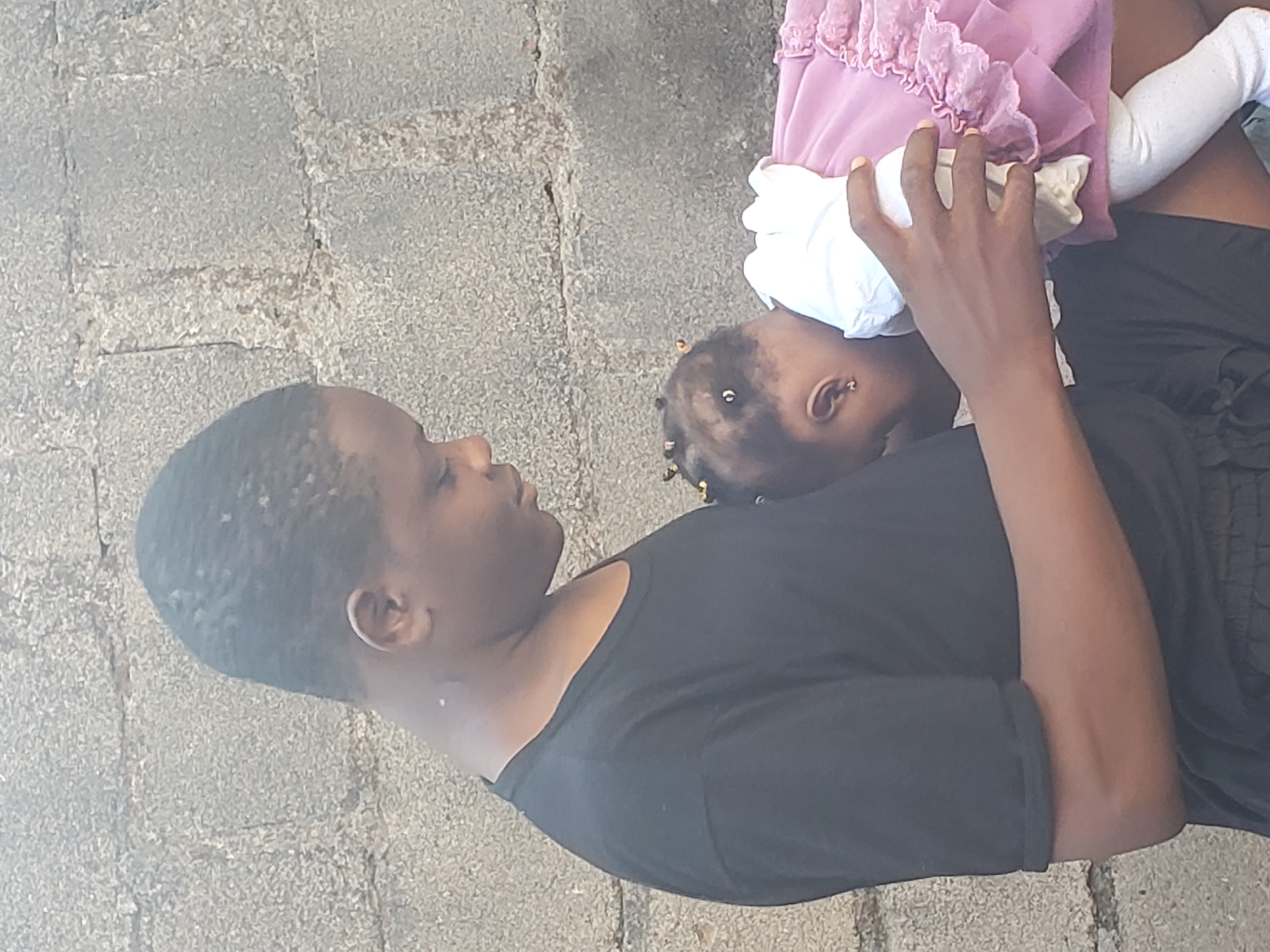 An adult and a young child are captured in a close, tender moment, appearing to be outdoors in Jos, Nigeria. The image shows the adult, likely a woman, lying or reclining on their side on the left, looking towards the child. They are wearing a simple black short-sleeved top. The child, a baby or toddler, is positioned on the right, nestled close to the adult. The child is dressed in a light pink ruffled top or dress and white leggings or socks.
The adult's right hand is gently placed near the child's head, appearing to hold or interact with a crumpled white cloth or bib, which is also close to the child's face. The child's face is partly visible, turned towards the adult, suggesting a moment of comfort, feeding, or close interaction. The child has small braids or beads in their hair and an earring visible in one ear.
They are situated on a paved surface made of irregularly shaped, light grey, textured stones or concrete blocks, indicating an outdoor setting. The lighting is bright and even, suggesting it is daytime. No other objects or visible text are discernible in the frame. The overall scene conveys a feeling of warmth, care, and connection between the adult and child.