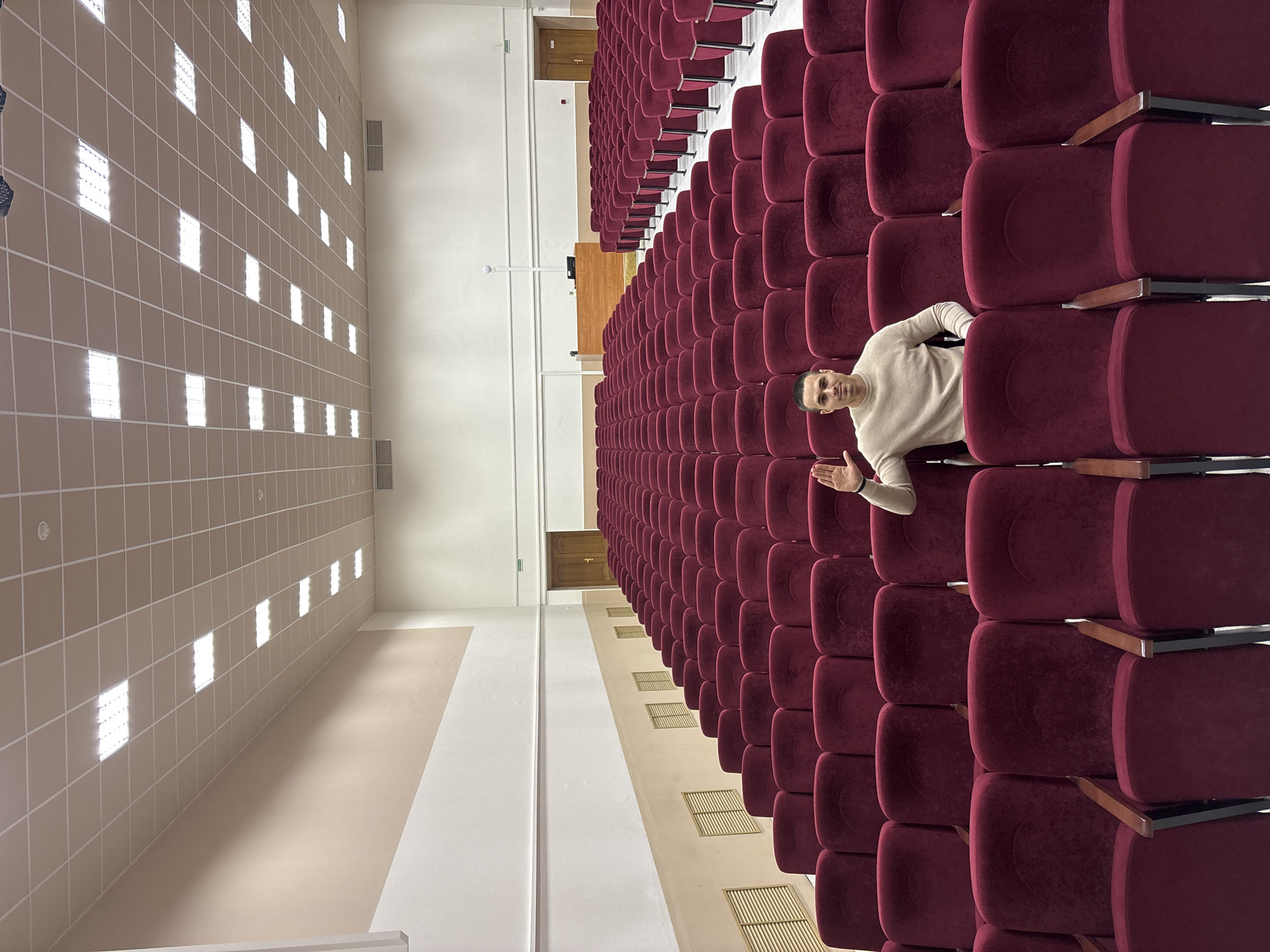A man stands in an auditorium in the city of Chaikovsky, Russia. The auditorium features rows of maroon-colored upholstered seating. The man, wearing a light-colored sweater, is positioned within the seating rows, facing the viewer. One arm is extended. Overhead is a grid of ceiling lights. A hallway is visible on the left side of the frame.