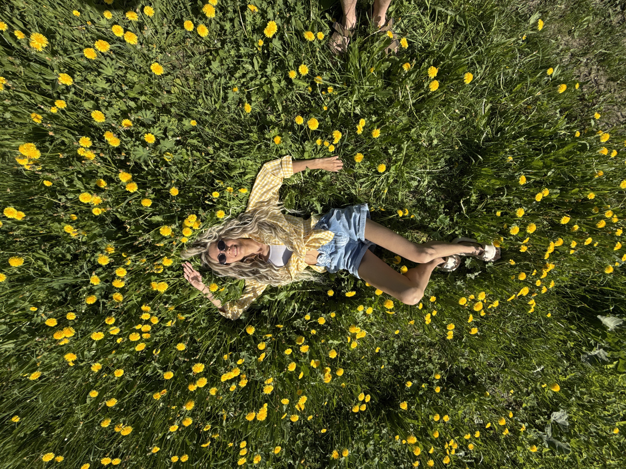 A woman lies on a grassy field covered with yellow flowers. She wears a yellow and white checkered shirt, blue denim shorts, sunglasses, and white shoes. Her blonde hair is spread out on the grass. Another person's feet are visible at the top of the frame. The scene appears to be outdoors during the daytime. The location is Шебунино, Russia.