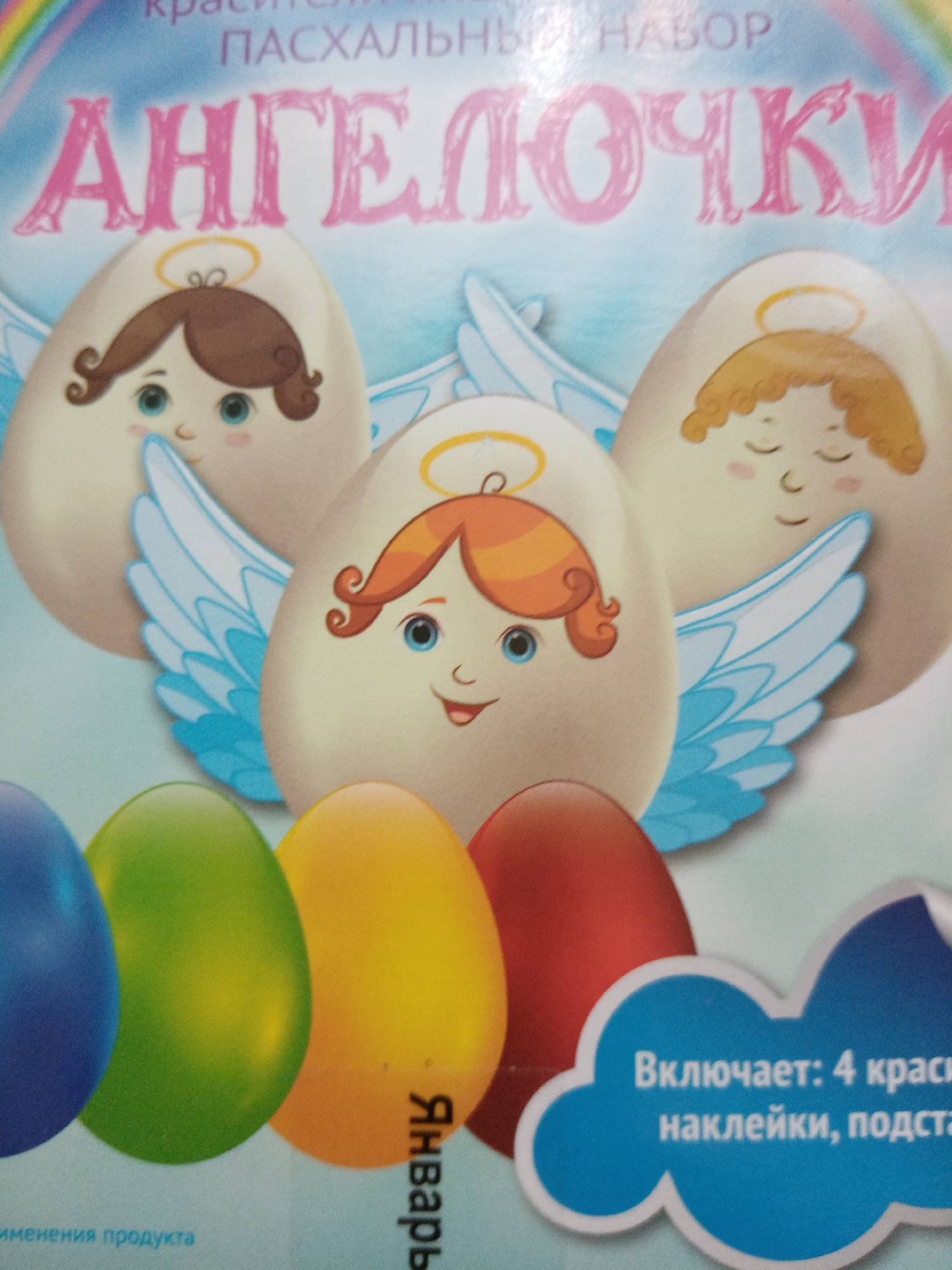 The image shows a close-up of a box for an Easter egg decorating kit labeled "Ангелочки" (Angels) in pink Cyrillic letters. The box features cartoon illustrations of three egg-shaped angels with halos and wings. Beneath the angels are four colored eggs: blue, green, yellow, and red. The word "Январь" (January) is visible on the yellow egg. A blue speech bubble indicates the contents of the kit: "Включает: 4 краси" followed by text continuing on a second line, "наклейки, подста". The box is photographed in Elektrostal, Russia.