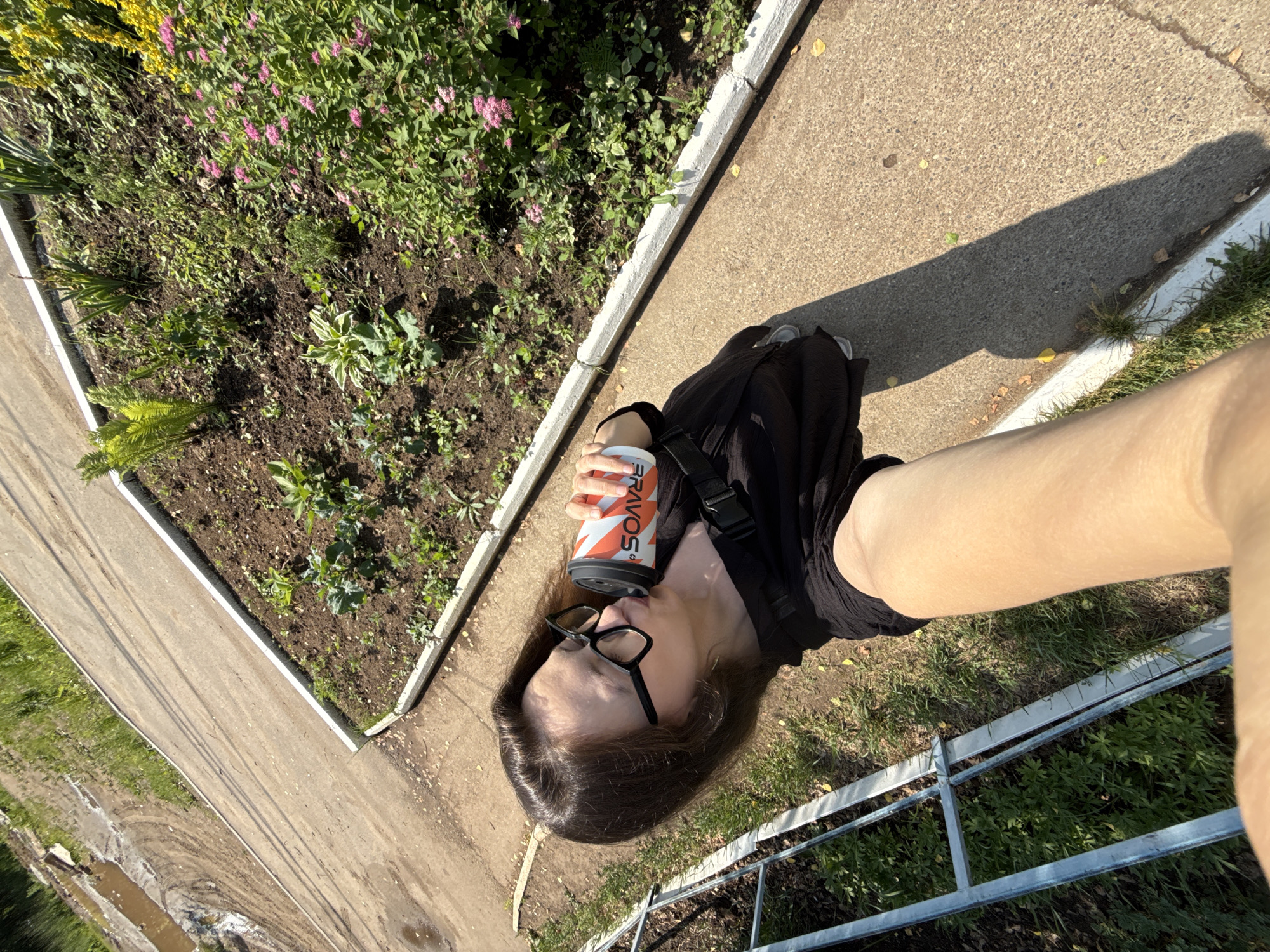 A person is taking a selfie outdoors, holding a cup of coffee and drinking from it. They are wearing a black top and glasses. The person is standing on a paved path next to a flower bed. The background shows grass, a fence, and a dirt path with puddles of water. The image appears to have been taken on a sunny day. The location is in Chaikovsky, Russia.