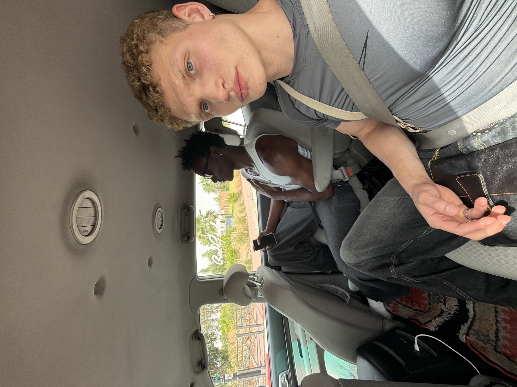 Two young men are seated in the back of a car, traveling through Mechouar Kasbah, Morocco. The man in the foreground, with light skin and curly blonde hair, is looking directly at the camera. He is wearing a grey t-shirt and a seatbelt. The man behind him, with dark skin and dark curly hair, is looking out the window. He is wearing a white tank top and grey sweatpants, and is holding a black smartphone. The car's interior is visible, including the headrests, air vents, and a small patterned rug on the floor. Outside the car, palm trees and greenery are visible, along with a sign that reads "Riad Garden". The overall impression is of a casual journey on a sunny day.