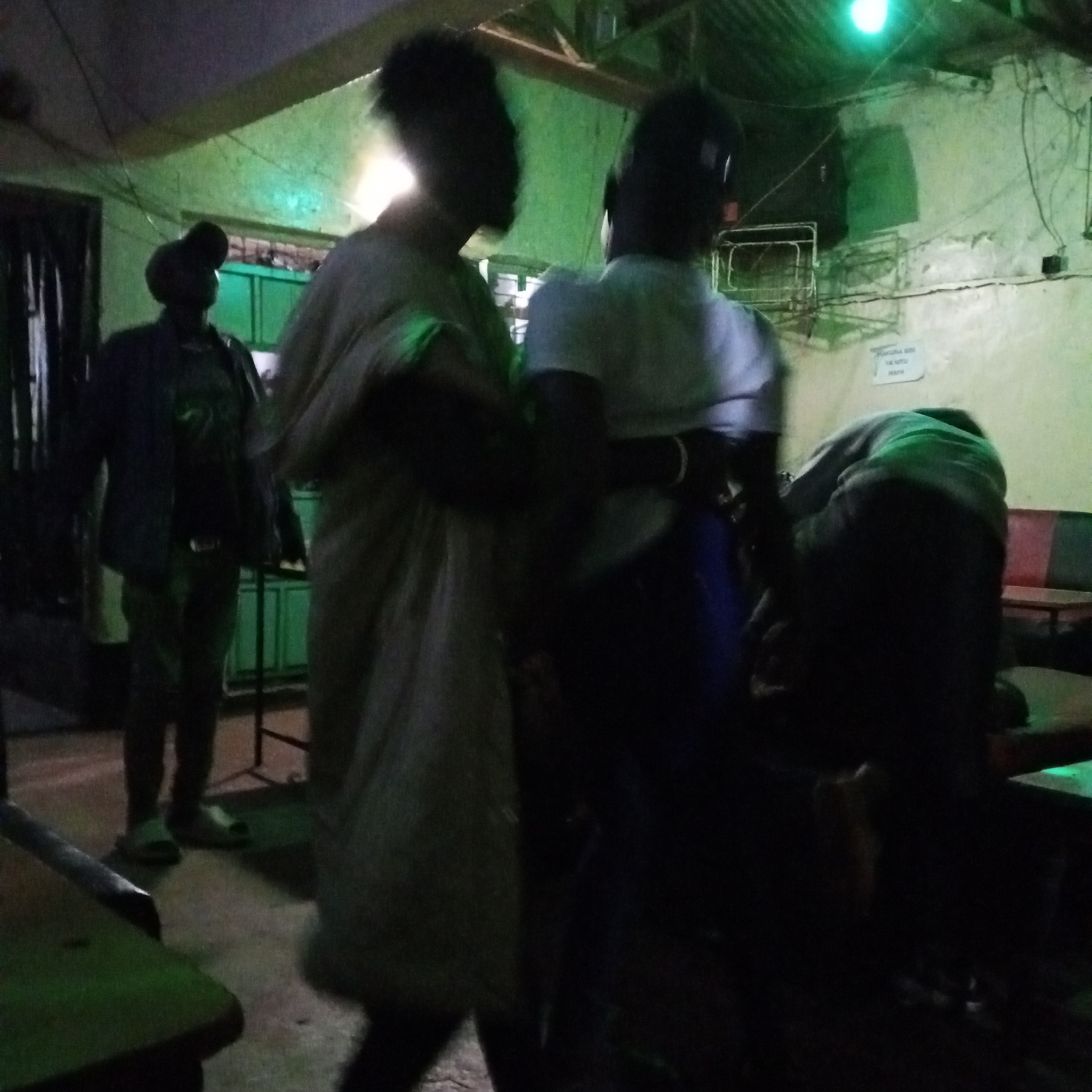 The image shows the interior of a dimly lit establishment in Borabu Chitago ward, Kenya. The lighting appears to be provided by a green-colored light source. The walls are pale and have visible wiring. There are at least four people in the shot. One person stands in the background wearing a jacket, pants, and a hat. Two people are partially visible in the foreground. One is wearing a light-colored garment. The other person is wearing a white shirt and dark pants. A person is bent over a table. Seating is also visible.