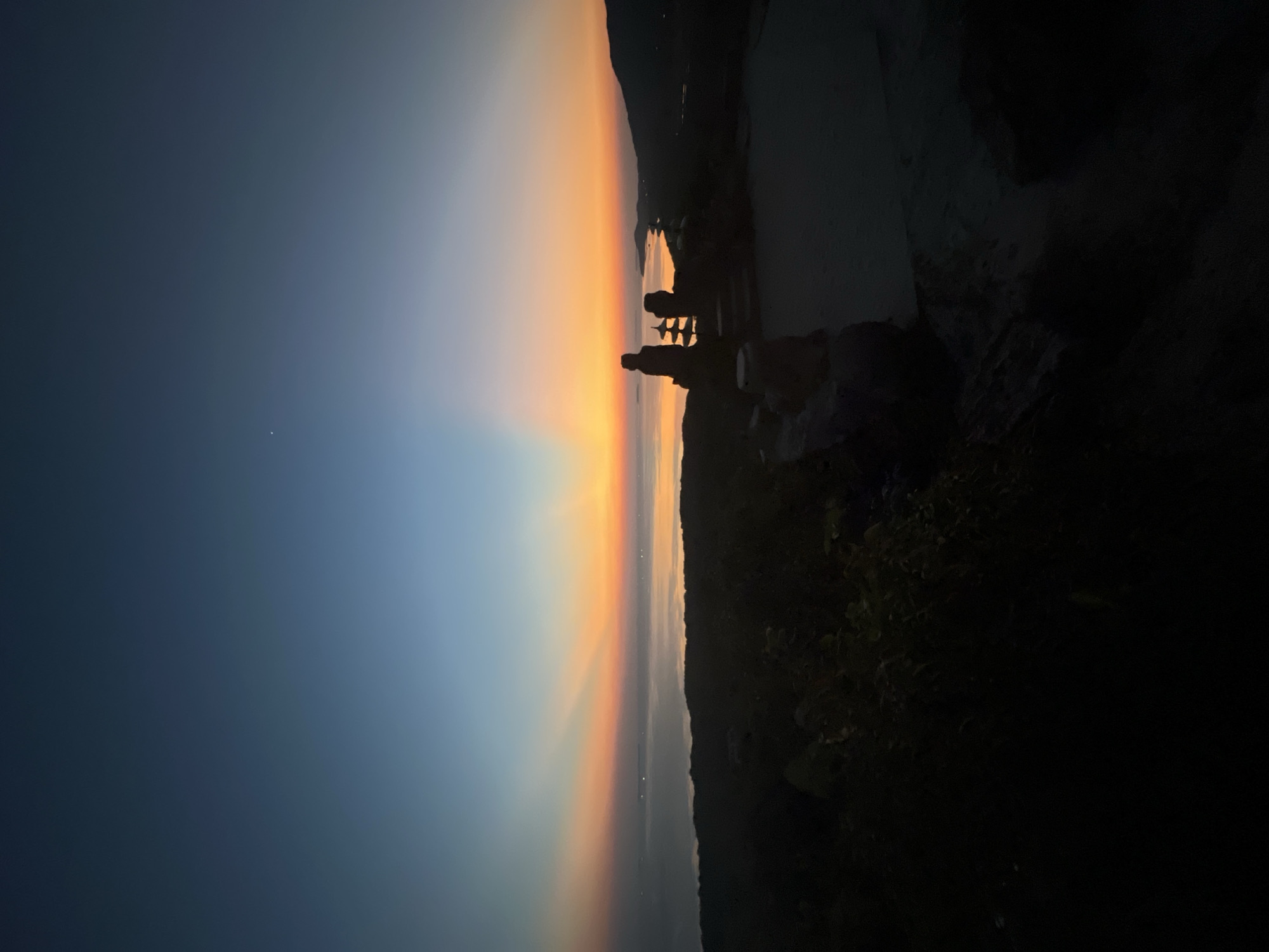 The image depicts a silhouetted landscape at sunset. The foreground features dark, shadowy foliage and a stone structure that resembles a small statue or monument. Beyond this, a body of water, likely the sea, is visible, with distant lights suggesting a coastal settlement or scattered boats. The sky transitions from a deep blue at the top to vibrant hues of orange, yellow, and pink near the horizon, indicative of a sunset. Rays of light fan out from the setting sun. The overall scene evokes a sense of tranquility and natural beauty. The location is identified as Karimunjawa, Indonesia. No text is visible in the image.