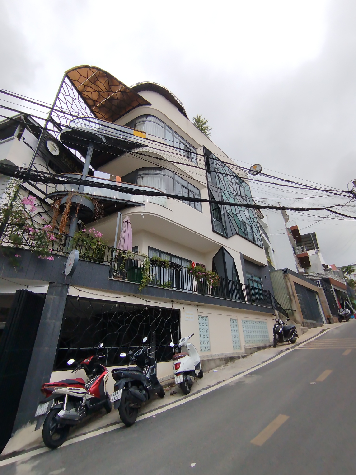 The image depicts a modern building with curved architectural elements located on a sloping street in Lam Vien - Da Lat, Vietnam. The building is multi-storied, primarily white, with black accents, a unique glass facade, and balconies with railings. Parked along the curb are several scooters. Overhead, power lines crisscross the sky. The overall environment appears to be a moderately dense urban area under a cloudy sky.