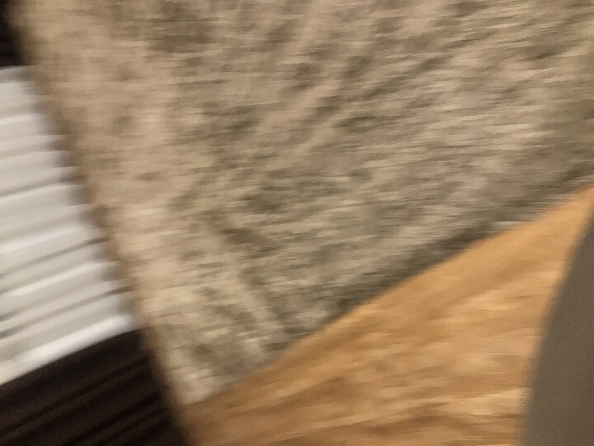 The image is blurry and lacks sharp details. I can discern the vague outline of a white, slatted object in the upper-left corner, possibly part of a window covering or furniture. Adjacent to it, there is a beige and brown patterned surface, perhaps a carpet or textile. A tan-colored, blurry area occupies the lower right, possibly a wooden floor or part of furniture. The lack of clarity prevents a definitive identification of objects or location. There is no indication of the city Yerevan, Armenia.