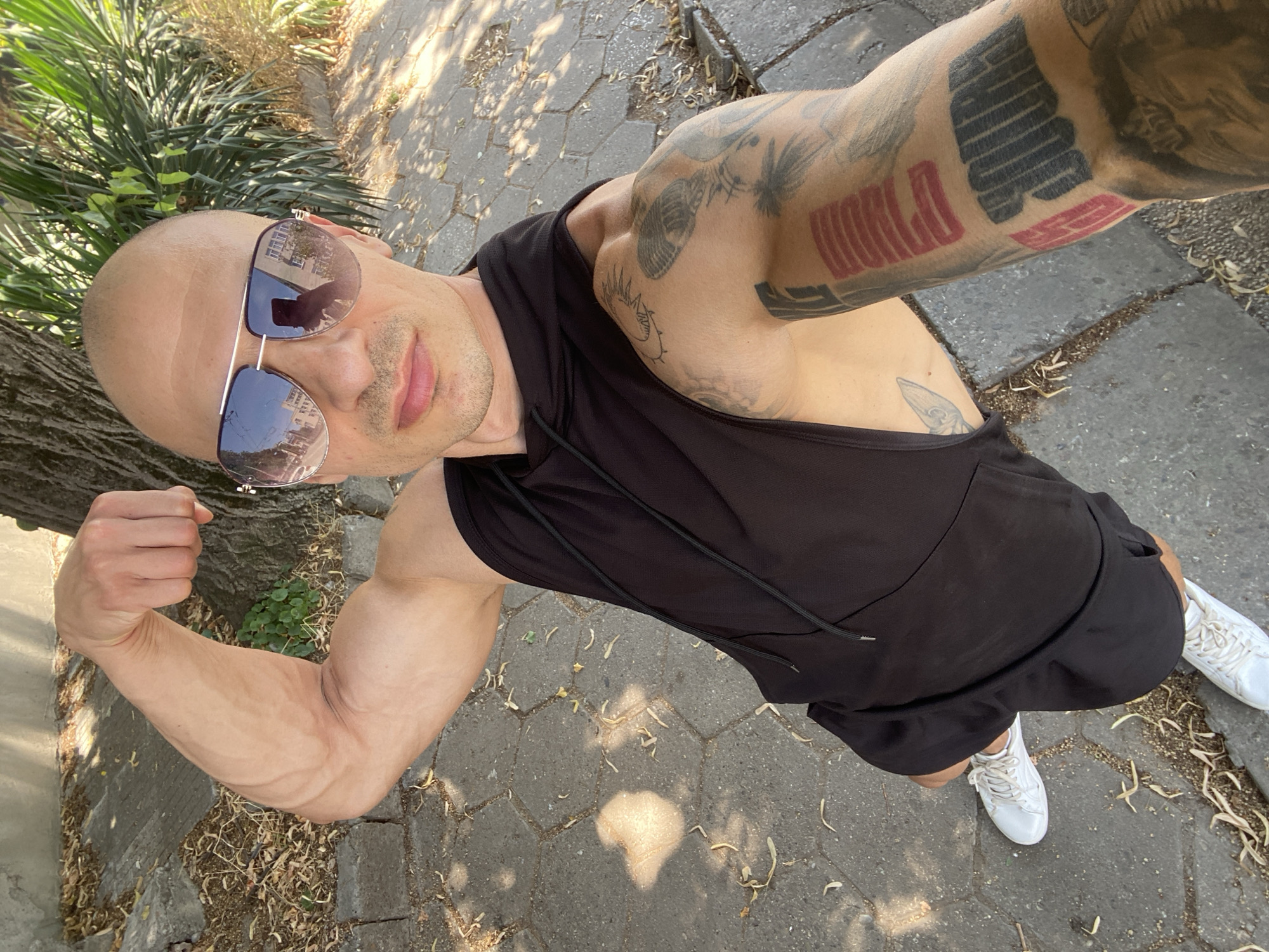The image shows a bald man with sunglasses and tattoos on his arms. He is wearing a black sleeveless hoodie and black shorts, with white sneakers. He is flexing his bicep, and his body is tanned. The background is an outdoor setting with a paved walkway, a tree on the left, and some greenery. Sunlight is filtering through the leaves of the tree, casting shadows on the ground. The man appears to be in a good mood, possibly posing for a photo. The time of day appears to be daytime. There is no visible text in the image. The provided location context is Pleven, Bulgaria.