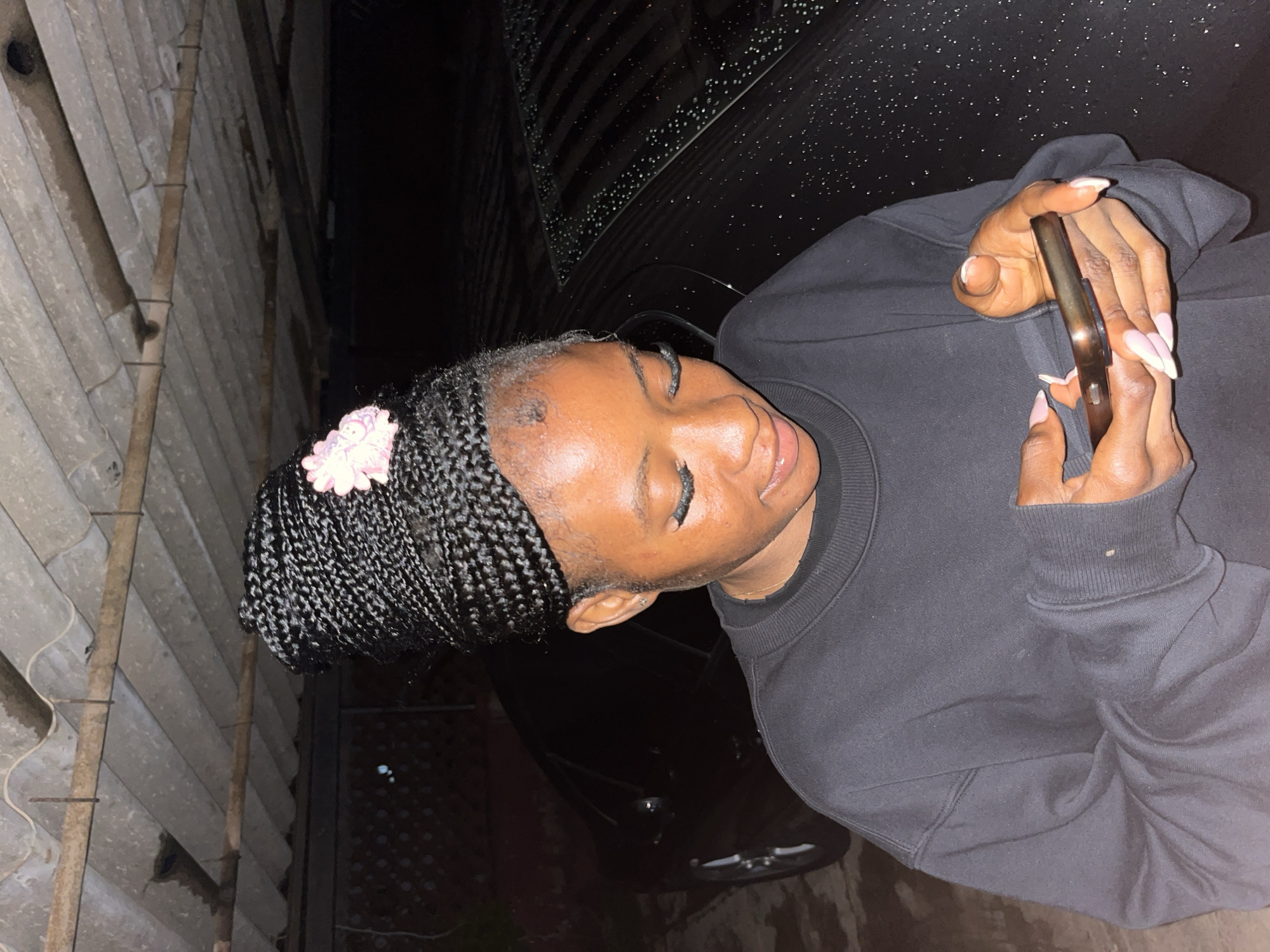 A dark-skinned young woman with braided hair adorned with a pink flower accessory is looking down at a smartphone she is holding with both hands. She is wearing a dark crew neck sweatshirt. The background shows a black car with water droplets on its surface, and a corrugated roof structure above. The scene appears to be outdoors, possibly in a covered parking area or carport, given the roof structure and the presence of a car. The lighting suggests it is evening or nighttime. The overall environment is dark and somewhat rustic.