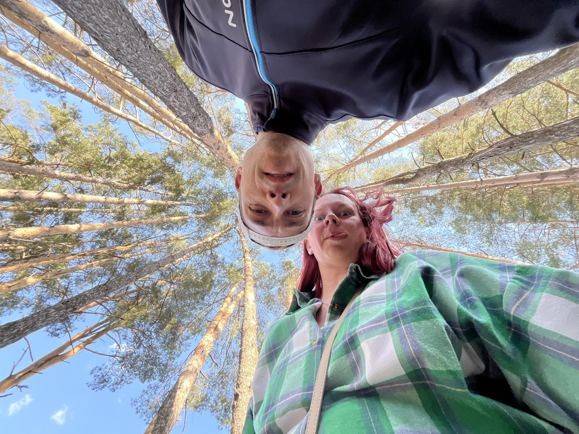 Two individuals are seen from a low-angle perspective looking down at the camera. The person at the top, presumed to be male, has a close-cropped haircut and wears a dark jacket with a blue zipper. The other individual, presumed to be female, has red hair and wears a green plaid shirt.
The background consists of a canopy of tall trees with a clear blue sky visible through the branches. Based on the location of Chaikovsky, Russia, it is likely a forest or park setting. The trees appear to be pine or similar conifer species.