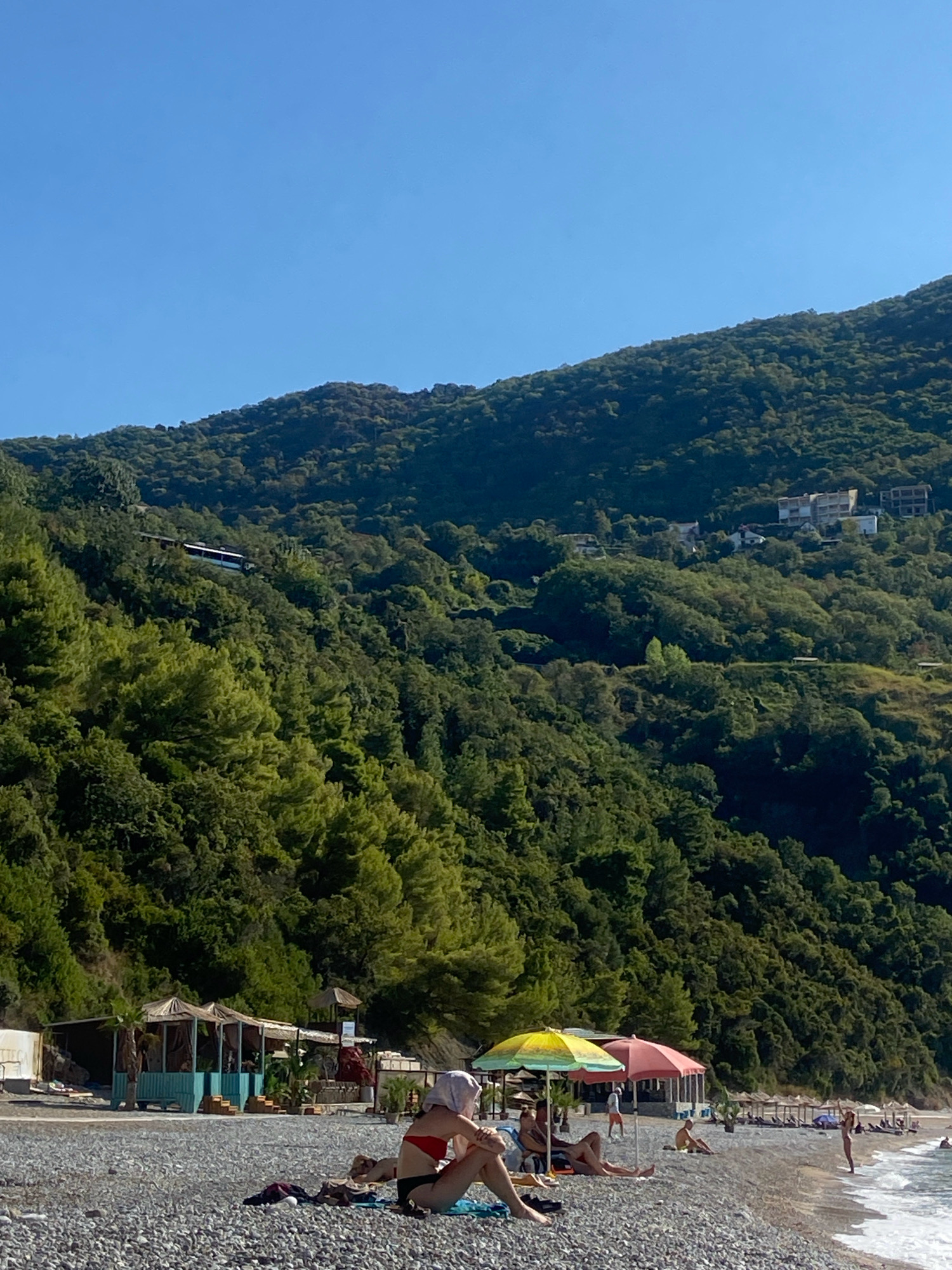 This image depicts a pebble beach adjacent to a dense, green, mountainous landscape. The sky is clear and blue. In the foreground, several individuals are seated or lounging on the beach. One person in the center foreground is wearing a red bikini and a white headscarf, sitting with their legs crossed. To their left, another person is visible, partially obscured.
Further down the beach, there are several colorful beach umbrellas, including a yellow and a pink one. Behind the umbrellas, a structure with open sides and a thatched roof appears to be a small beachside cafe or bar, with tables and chairs visible. Beyond this, rows of empty sun loungers and umbrellas are set up along the shoreline.
The mountainous backdrop is covered in a thick forest of various shades of green trees. A road or path is visible winding up the side of the mountain, with a long, white vehicle, possibly a bus, traveling on it. Higher up on the slope, several buildings, including what appear to be apartment blocks or villas, are situated amongst the trees. The sea is visible on the right side of the image, with gentle waves lapping at the shore. The scene suggests a tranquil day at a coastal resort area, likely in Prijevor, Montenegro, given the characteristic mountainous coastline.