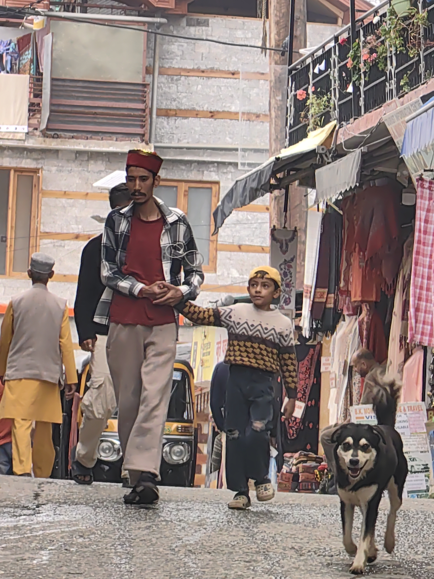 In Vashisht, India, a narrow, paved street slopes downwards, lined with buildings. On the left, a man in a red cap and checkered shirt walks forward, his hands clasped. A young boy wearing a yellow cap and a patterned sweater walks slightly ahead of and to the right of the man, his left hand extended. Further to the left, another man in a yellow kurta and grey vest walks away from the camera. Behind the man in the checkered shirt, a yellow auto-rickshaw is visible. On the right side of the street, market stalls display various textiles. In the foreground, a medium-sized dog with black and white markings trots towards the camera, its mouth open and tongue visible. The overall scene depicts a busy street in a village setting.