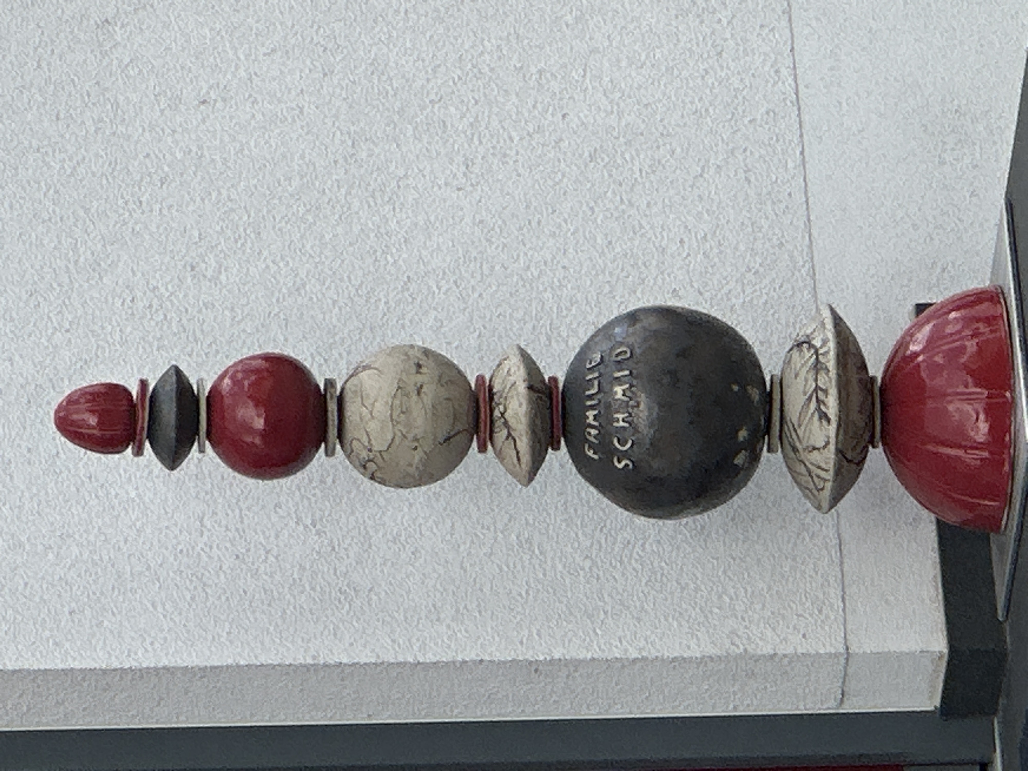 The image shows a tall, abstract sculpture made of stacked, rounded elements in various colors and textures. The sculpture is positioned in front of a textured white wall, which appears to be the exterior of a building. On the left side of the image, a dark gray vertical element, possibly a window frame or door frame, is partially visible. The sculpture consists of a red, egg-shaped top, followed by a dark gray flattened disc, a red sphere, a light-colored, cracked sphere, a red disc, another light-colored, cracked, flattened oval shape, a large dark gray sphere with the inscription "FAMILIE SCHMID" in gold lettering, another light-colored, cracked, flattened oval shape, and a large red dome at the base. The sculpture is situated outdoors, and the lighting suggests it is daytime. This sculpture is located in Günzburg, Germany, at the entrance of the "Familie Schmid" bakery.