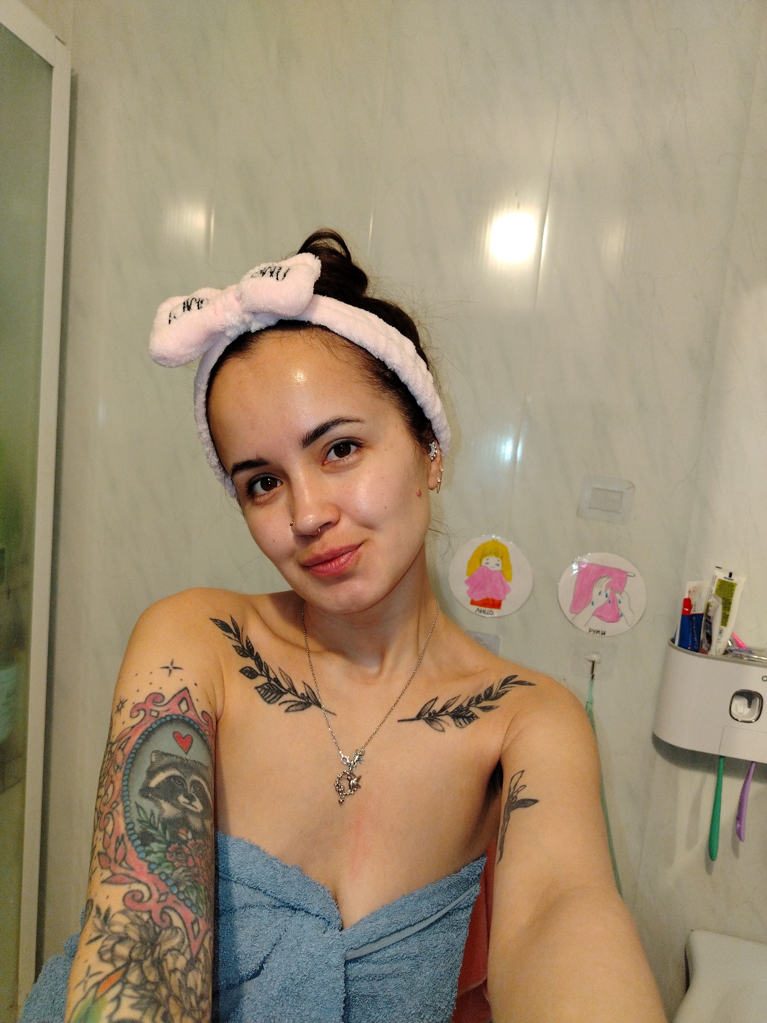 A woman with a pink headband stands in a bathroom. She has tattoos on her arms and chest. She is wearing a blue towel. On the wall are two drawings, a toothbrush holder, and tiles. The location is Noviy, Russia.