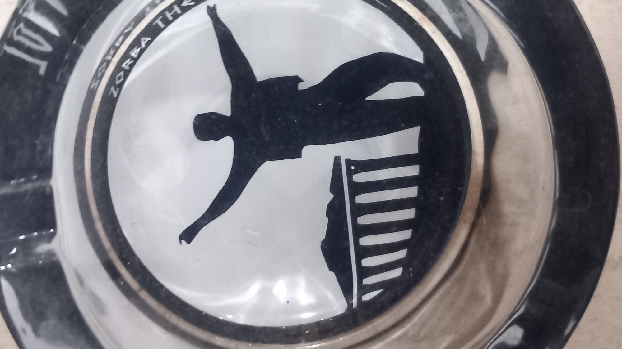 This is a close-up, slightly angled shot of a clear glass ashtray with a black and white graphic design. The graphic depicts a silhouetted male figure with arms outstretched, standing next to a row of white columns. The figure appears to be dancing or in mid-movement. Text is visible around the inner rim of the ashtray; the visible words include "SORRY" and "ZORBA TH". The ashtray itself has a thick black rim and a silver band encircling the graphic. The background appears to be a light-colored, textured surface. This ashtray is likely a souvenir from Kos, Greece, referencing the famous novel and film "Zorba the Greek."