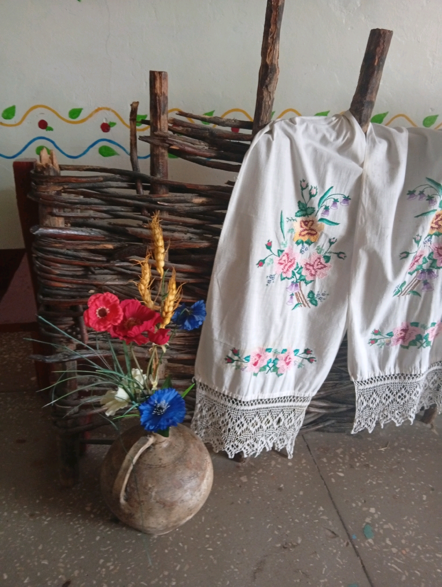 A still life composition is presented in an indoor setting with a white wall adorned with a colorful painted border. Dominating the scene is a woven basket made of dark branches, which serves as a backdrop for a bouquet of artificial flowers. The bouquet features red poppies, blue cornflowers, wheat stalks, and some white flowers, all arranged in a rustic, round, earthen pot. To the right of the basket, a white embroidered fabric hangs, draped over wooden posts. The fabric is decorated with floral embroidery in shades of pink, yellow, green, and purple, and features a lace trim along its bottom edge. The floor is concrete, with a speckled pattern of light and dark grey. The overall impression is one of folk art and rural aesthetics.