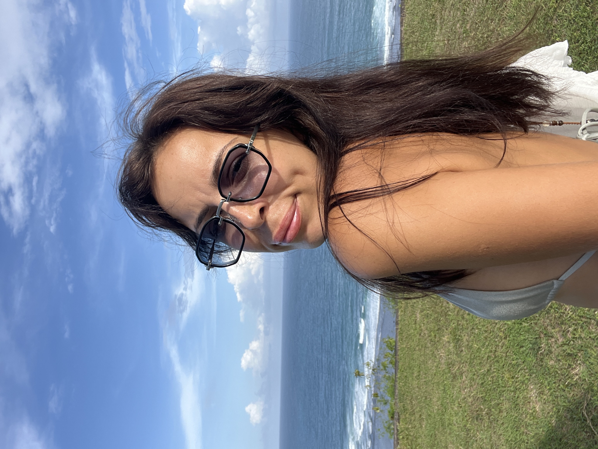 A young woman is captured in a close-up, outdoor portrait set against a scenic coastal backdrop, likely in Kuta Selatan, Indonesia. She is looking towards the viewer with a slight, contented smile, wearing stylish, dark sunglasses with uniquely shaped frames. Her long, dark hair flows around her, and she appears to be dressed in swimwear, with a light grey bikini strap visible.
The setting features a bright blue sky dotted with scattered white clouds above a vast, calm blue ocean where gentle waves are breaking near the shore. A strip of vibrant green grass, indicating a cliff edge or hillside, lies between the woman and the sea. The clear illumination suggests it is bright daylight, possibly midday or early afternoon, indicative of pleasant, sunny weather. The overall impression is one of leisure and enjoyment in a beautiful tropical environment. There is no visible text in the image.
