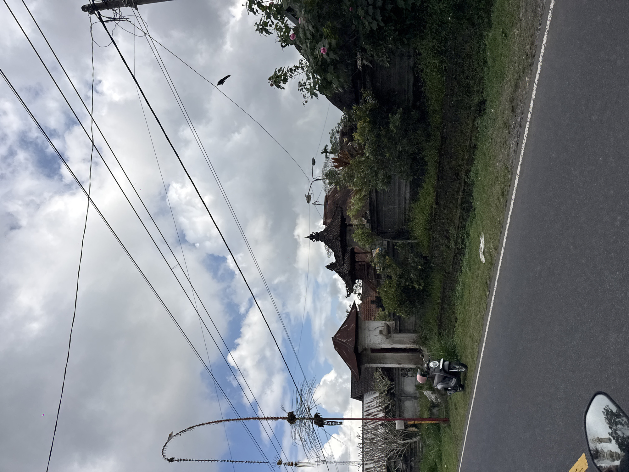 The image depicts a street scene in Susut, Indonesia, with a focus on a traditional Balinese structure and a scooter. The viewpoint is low, suggesting the photo was taken from a vehicle, possibly a motorcycle, given the presence of a rearview mirror in the lower right corner reflecting trees and a building.
The main subjects include a dark-colored scooter with a pink helmet parked on the grassy verge beside a paved road. Behind the scooter, a building with traditional Balinese architecture is visible. It features tiered, dark-tiled roofs and ornate detailing. The building appears to be partially overgrown with lush green vegetation, including flowering plants with pink blossoms.
In the foreground, a power pole with numerous overhead wires dominates the left side of the image. These wires are a prominent visual element, creating a network against the sky. A bird is silhouetted against the sky, perched on one of the wires. Further down, a decorative string of what appears to be fish bones or a similar material stretches across the frame, likely part of a local tradition or celebration.
The sky is partially cloudy, with patches of blue visible. The lighting suggests it is daytime, but the clouds might indicate the possibility of rain or overcast conditions. The overall impression is one of a rural or semi-rural environment, characterized by a blend of everyday infrastructure, traditional architecture, and abundant natural greenery. The image conveys a sense of a peaceful, everyday moment in a tropical setting. There is no visible text in the image.