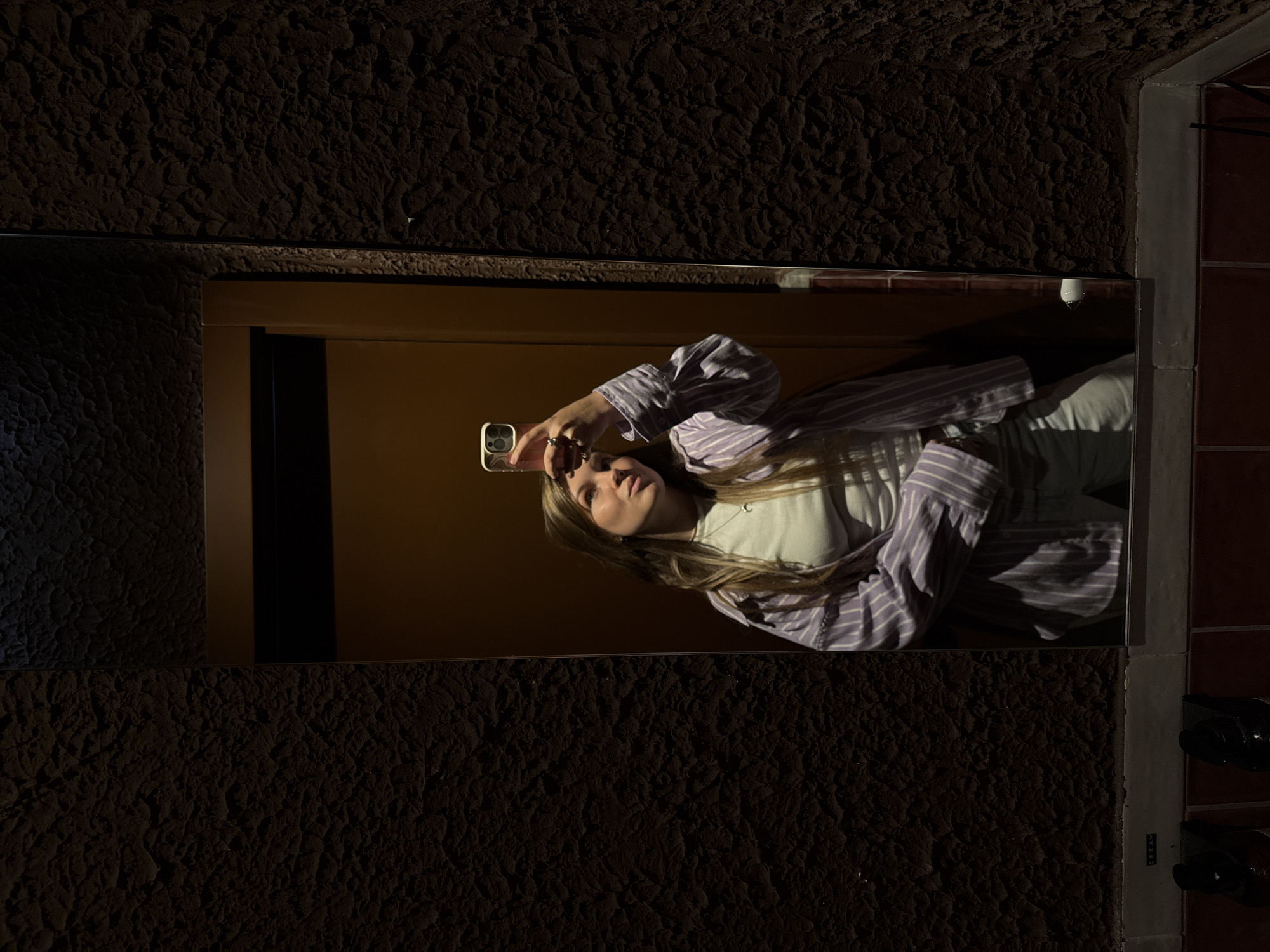 A blond woman, visible in a mirror's reflection, is taking a selfie with her phone. She is wearing a white t-shirt and a purple striped shirt. The mirror is mounted on a dark, textured wall. To the right of the mirror, there is a tiled section with a coat rack holding dark shoes. The reflection in the mirror shows a dark-yellow door. The location is Belgrade, Serbia.