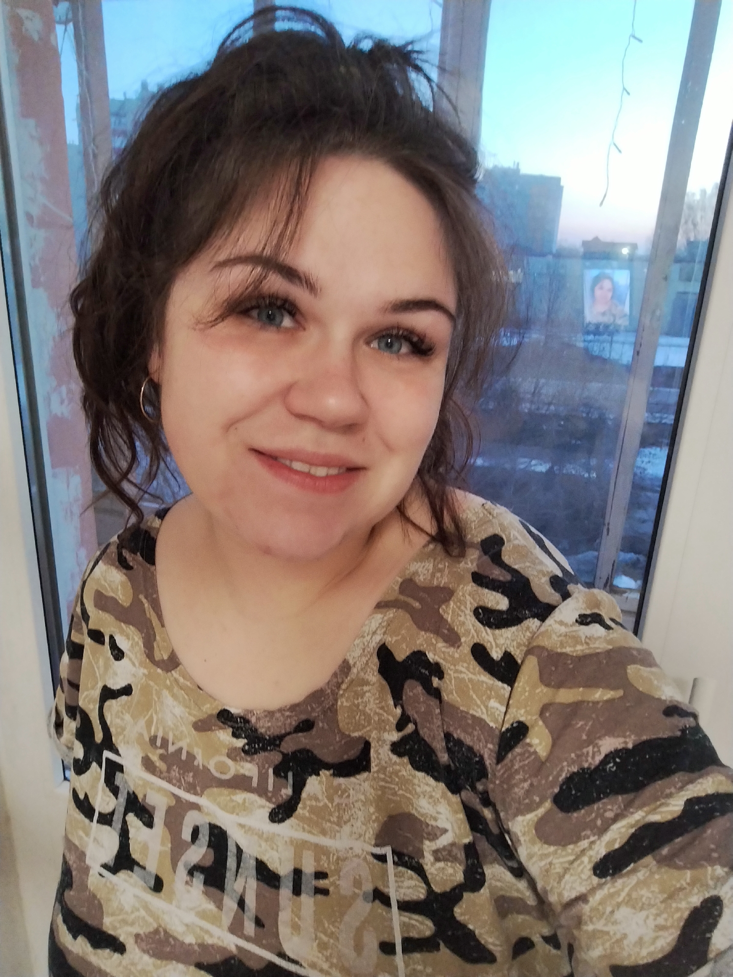 A woman with dark hair pulled back, blue eyes, and a camouflage-patterned shirt is smiling into the camera. She is wearing a gold hoop earring in her left ear. She is standing in front of a window, and outside the window, a city scene is visible. There is a building with a picture of a woman on its side. Above the picture, there appear to be some hanging lights. The city is in Chaikovsky, Russia.