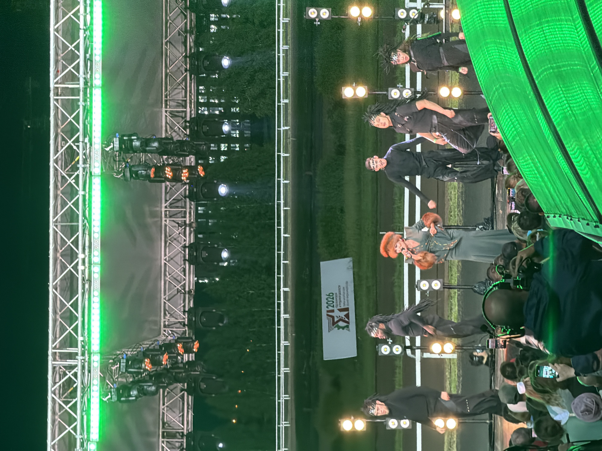 In an outdoor setting, a group of performers is on stage with a backdrop of trees and a horse racing track visible behind them. The stage is illuminated by green lights. In the foreground, a large audience is gathered, many holding up phones. A person in the audience is wearing a blue jacket and headphones, appearing to be filming or recording. The performers on stage are dressed in dark, theatrical costumes, some with elaborate headdresses. One female performer in the center is wearing a green dress and has bright red hair. A banner with the text "12026" and a logo is displayed on the stage. The overall scene suggests an outdoor concert or performance event.