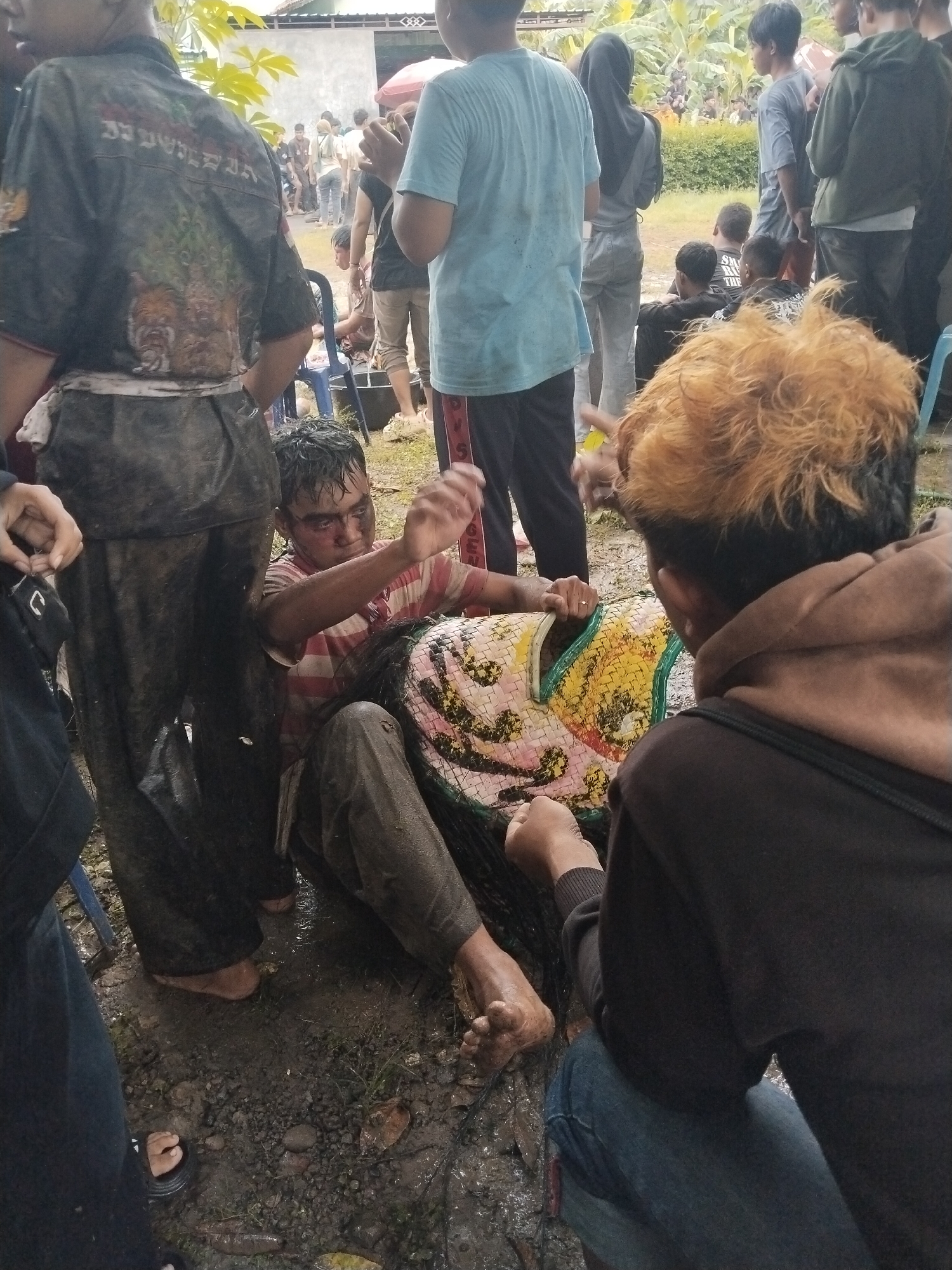 Location: Karang Tengah, Indonesia.

A group of people are gathered in an outdoor area with a wet, muddy ground. Several individuals are standing, while one man is seated on the ground, appearing to be involved with a large, colorful mask. Two other individuals are close to him, seemingly assisting or interacting with the mask as well. The crowd around the central group suggests a gathering or event. Mud is splattered on the clothes of the seated man and at least one of the standing observers. The general atmosphere appears to be overcast.