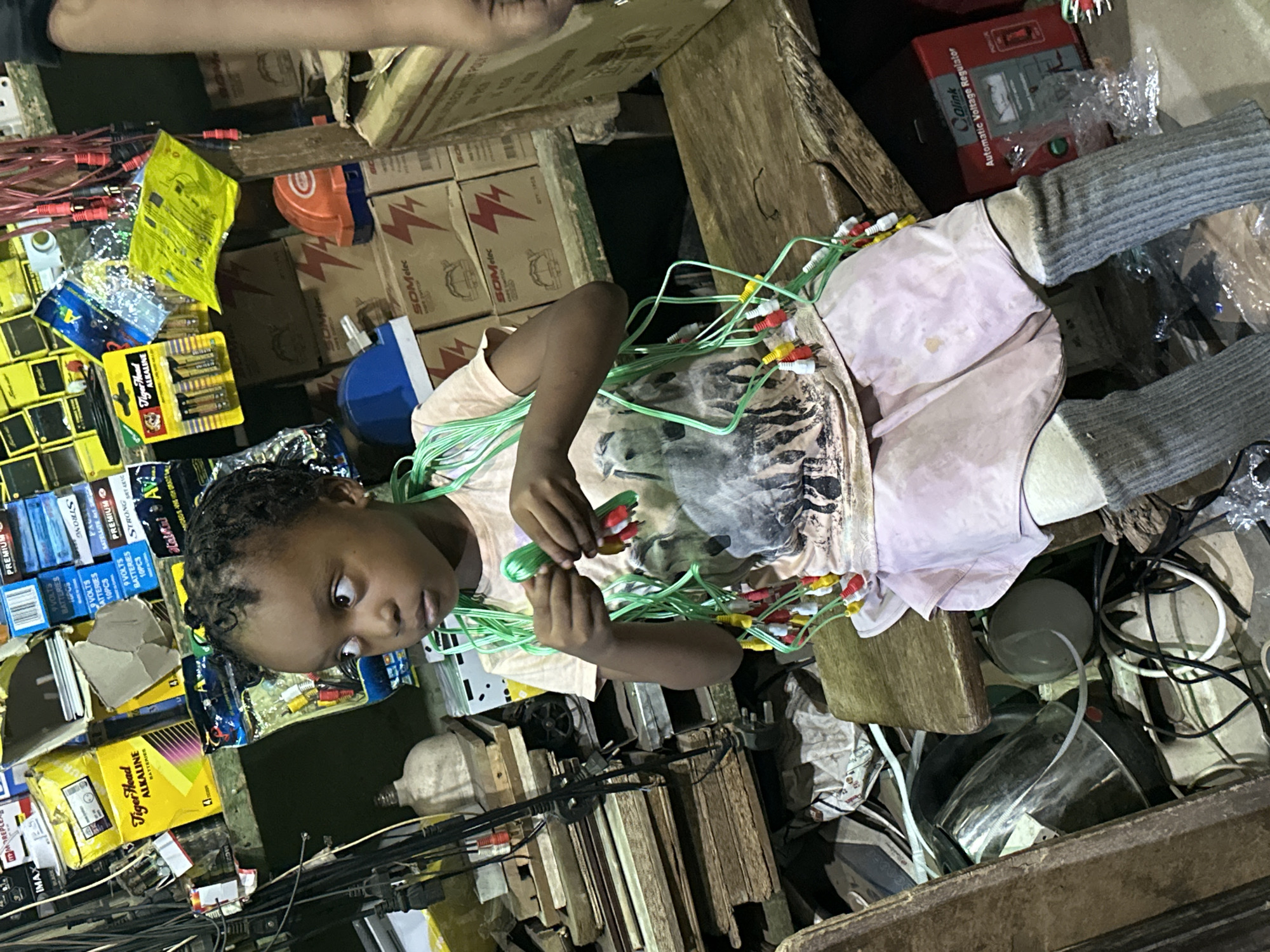 In Barden Yana, Nigeria, a young child is engrossed in handling a bundle of brightly colored audio-visual cables, their ends adorned with red, yellow, and white connectors. The child, wearing a light-colored t-shirt with a graphic design and grey socks, appears focused on the task, possibly sorting or organizing the wires.
The setting is a cluttered indoor space, likely a market stall or a storage area, packed with various electronic components and packaged goods. Stacks of cardboard boxes, some featuring lightning bolt logos and the brand "SOMelec," are visible, alongside blister packs of "Tiger Head ALKALINE BATTERIES." Other items like an "Automatic Voltage Regulator" from "Olink" and numerous other battery boxes are also present, indicating a business dealing in electronics and related accessories.
The lighting suggests an indoor environment, possibly dimly lit or with artificial light sources. The overall scene conveys a sense of a busy marketplace or a workshop, with the child actively interacting with the products. The child's expression is one of concentration, with wide eyes directed at the cables. The surrounding chaos of wires and boxes hints at a dynamic environment where such items are stored and sold.