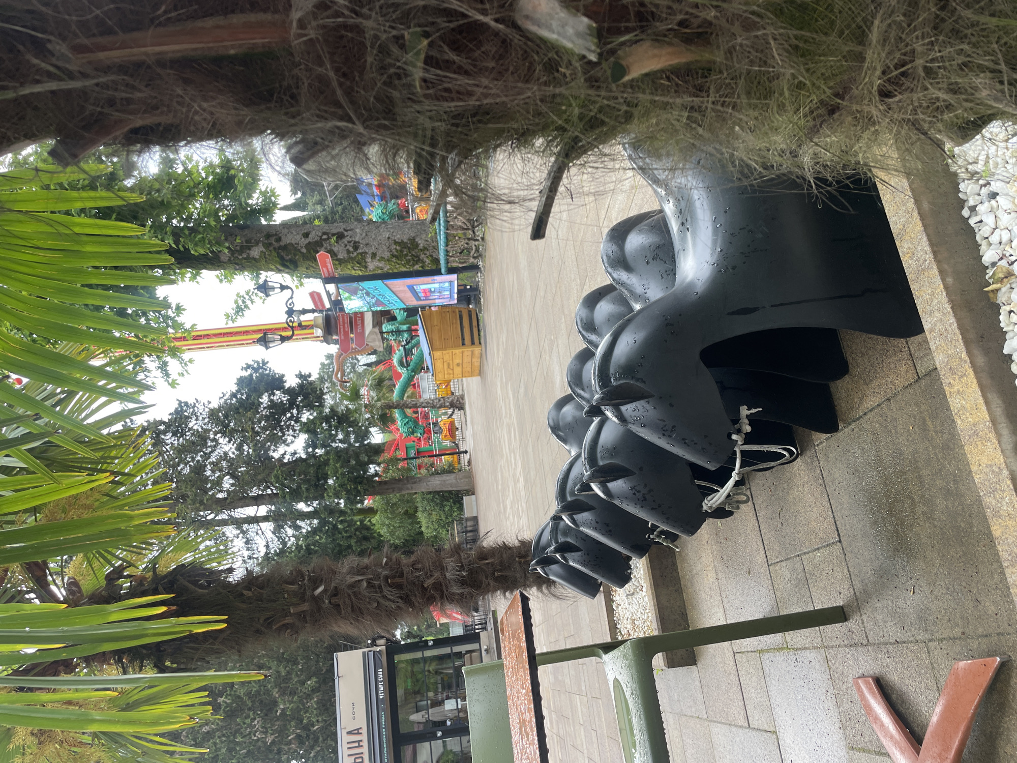 This outdoor shot, taken in Sochi, Russia, shows a row of black, plastic, watercraft-shaped vehicles. The vehicles are linked together with white rope and are situated on a paved area, likely a sidewalk or plaza. A low, olive-green metal railing or barrier is partially visible in the foreground.
In the background, through an opening created by palm tree trunks and foliage, one can see a distant amusement park ride, trees, and other unidentified structures. A sign with Cyrillic text, possibly indicating "ITALIAN PIZZA" and "CHETYRE SYNA SOCHI," is partially visible near the base of one of the palm trees. The scene appears to be taken on an overcast day, with water droplets visible on the vehicles.
