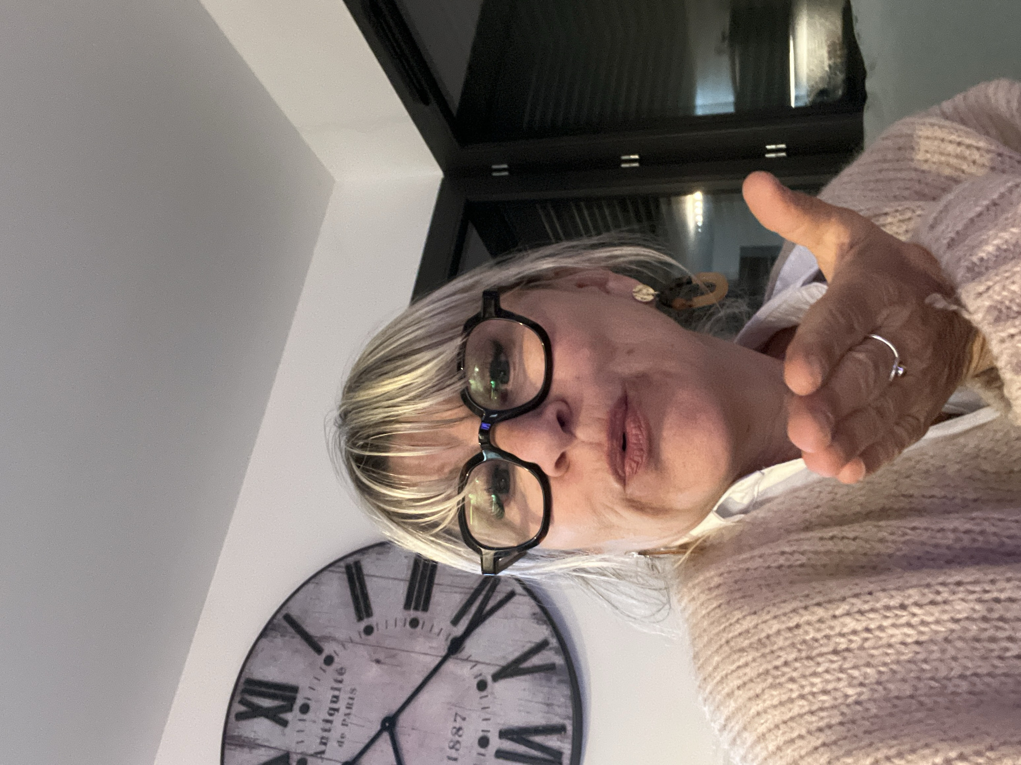 A woman with blonde hair and black-rimmed glasses stands indoors with a large clock on the wall behind her. The clock has Roman numerals and the words "Antiquité de Paris, 1887" printed on its face. The woman is wearing a light-colored knitted sweater. Her right hand is raised in a gesture. In the background, there is a dark window frame. It is not possible to confirm the location in the city of Chambœuf, France.