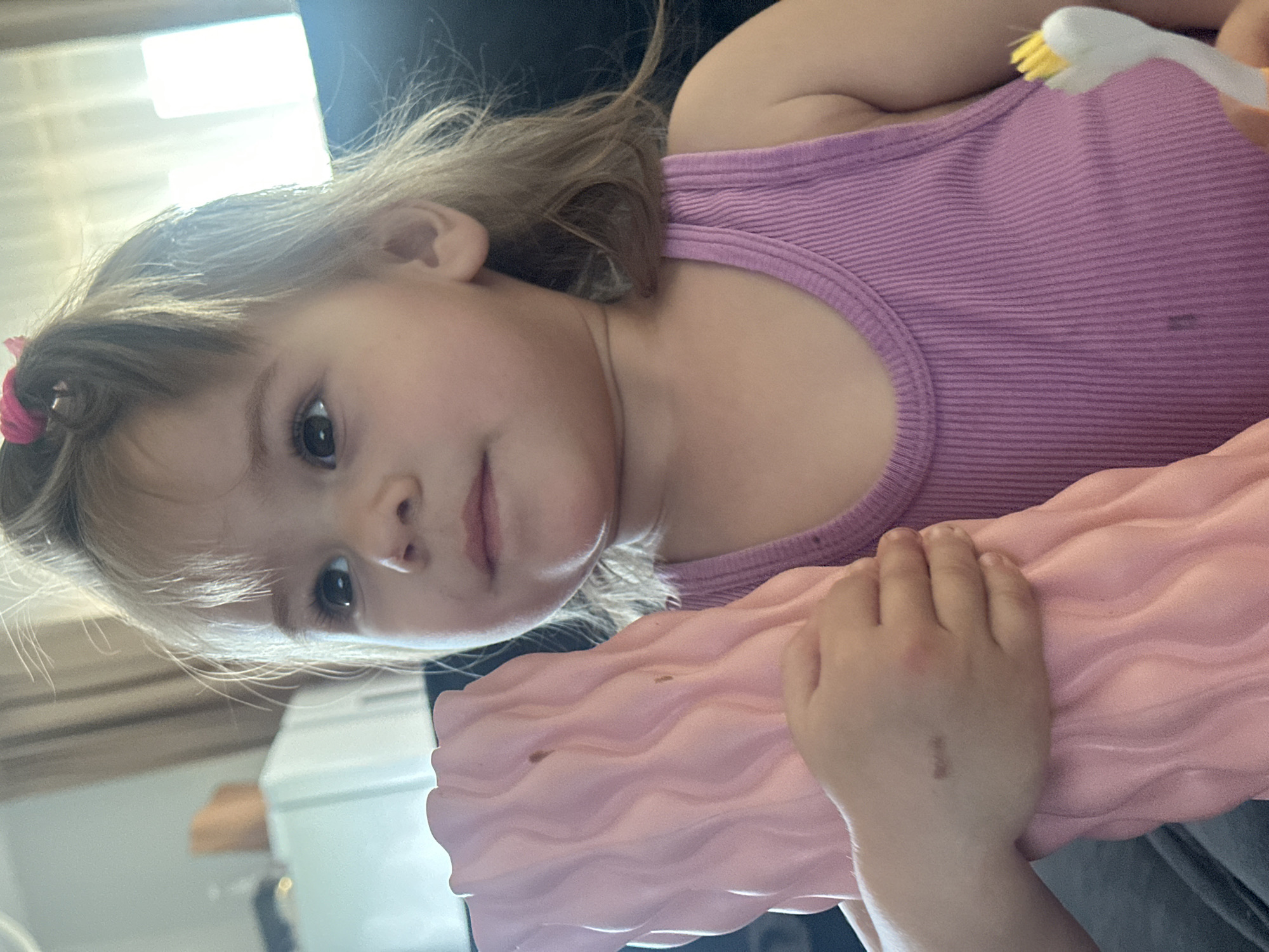 A young child, likely a girl with light brown hair and brown eyes, is depicted indoors, looking directly at the camera. The image is tilted, showing her seemingly reclining or lying on her side. She is wearing a ribbed, purple tank top with a couple of small, dark marks on it. A small pink hair tie or clip is visible in her hair on the left side of her head.
The child holds a large, pink, wavy-textured object, possibly a toy or cushion, with her right hand. Faint marks are visible on the back of her hand and on the pink object. In the upper right corner of the frame, a white toothbrush with yellow bristles is partially visible, held by an unseen hand. The background is softly lit, suggesting an indoor setting with natural light coming from a window or bright source in the upper left. A dark area, possibly furniture, is behind her head, and a blurry white object, perhaps an appliance or cabinet, is in the lower left background.
The child's expression is calm and neutral. The presence of the toothbrush suggests an activity related to oral hygiene or imaginative play. The indoor setting does not provide specific geographical cues to confirm the location as Kharnavy, Russia. There is no visible text in the image.