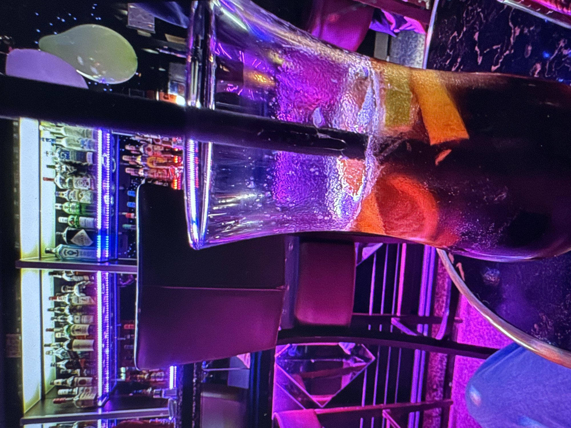 The image depicts a close-up view of a cocktail in a tall glass, with a dark beverage and slices of fruit such as oranges and possibly grapefruit. A black straw is visible in the drink. The setting appears to be a bar, with shelves stocked with numerous bottles of liquor and spirits in the background. The area is illuminated with vibrant purple and blue neon lights, creating a lively atmosphere. In the mid-ground, a dark-colored bar stool with a purple cushion is partially visible. Further in the background, there are balloons, one of which is light green and partially deflated. The image is captured from a slightly elevated angle, focusing on the drink and the immediate surroundings. There is no discernible indication of the location being in Porrentruy, Switzerland.