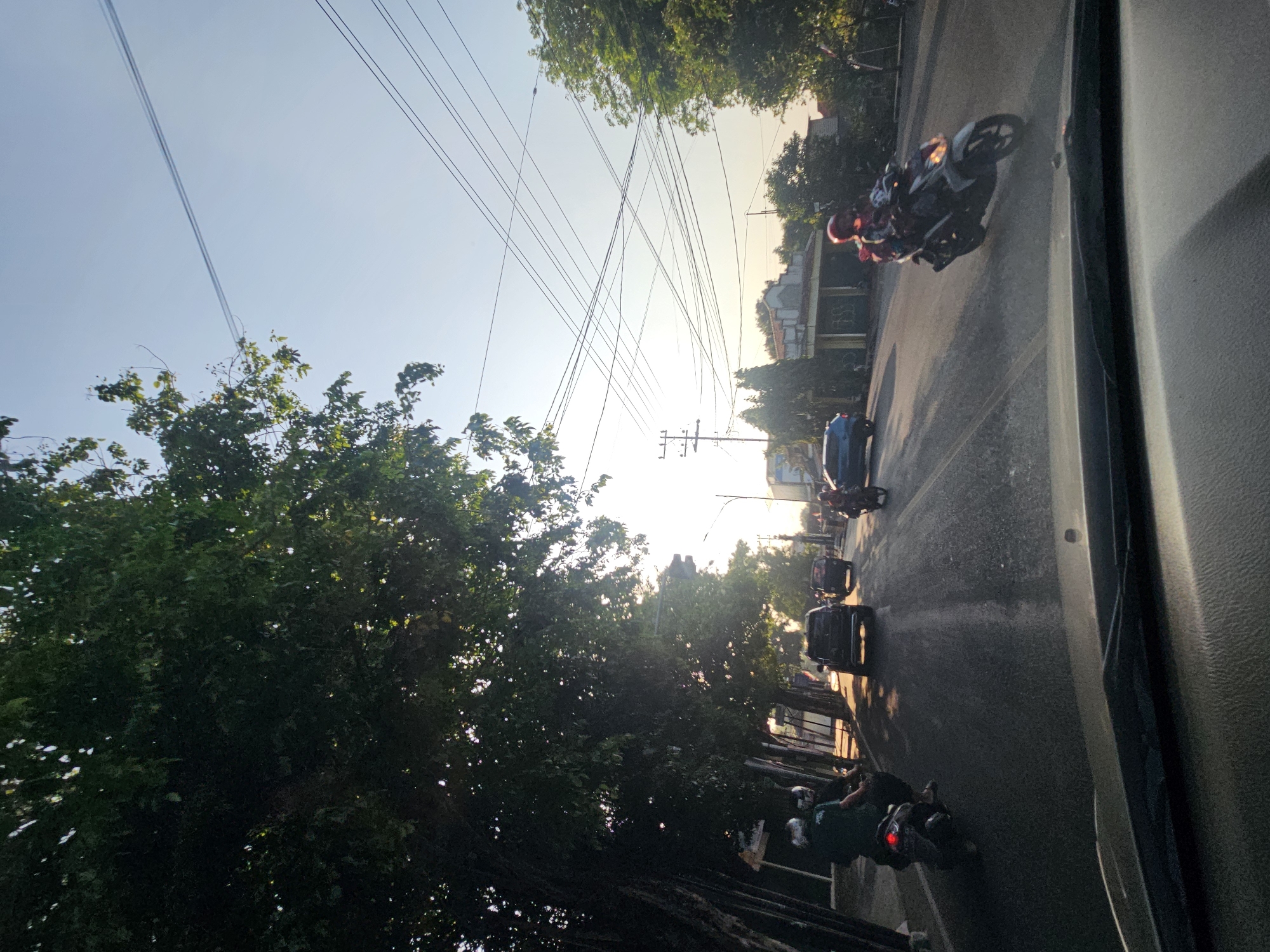 The image captures a street scene in Tuban, Indonesia, during what appears to be late afternoon or early morning with the sun low in the sky, casting long shadows and a warm glow. The perspective is from inside a vehicle, looking forward.
On the left side of the road, a dense canopy of trees with dark green leaves shades a sidewalk area. Underneath these trees, two individuals on a scooter are visible. The person in the back is wearing a dark green shirt and helmet, while the driver also wears a helmet.
The road itself is paved and appears to be a multi-lane street with moderate traffic. Several vehicles are present: a dark SUV is driving in the same direction, followed by another darker vehicle. Further ahead, a blue car is driving in the opposite lane. On the right side of the road, a motorcycle with a rider wearing a red helmet and jacket is moving towards the viewer.
The environment is urban, with buildings and power lines visible along the sides of the street. A utility pole with multiple wires is prominent in the middle distance. The sky is clear and light blue, with the sun creating a bright halo effect near the horizon. The overall impression is a typical day of transportation and daily life in a moderately busy Indonesian city.