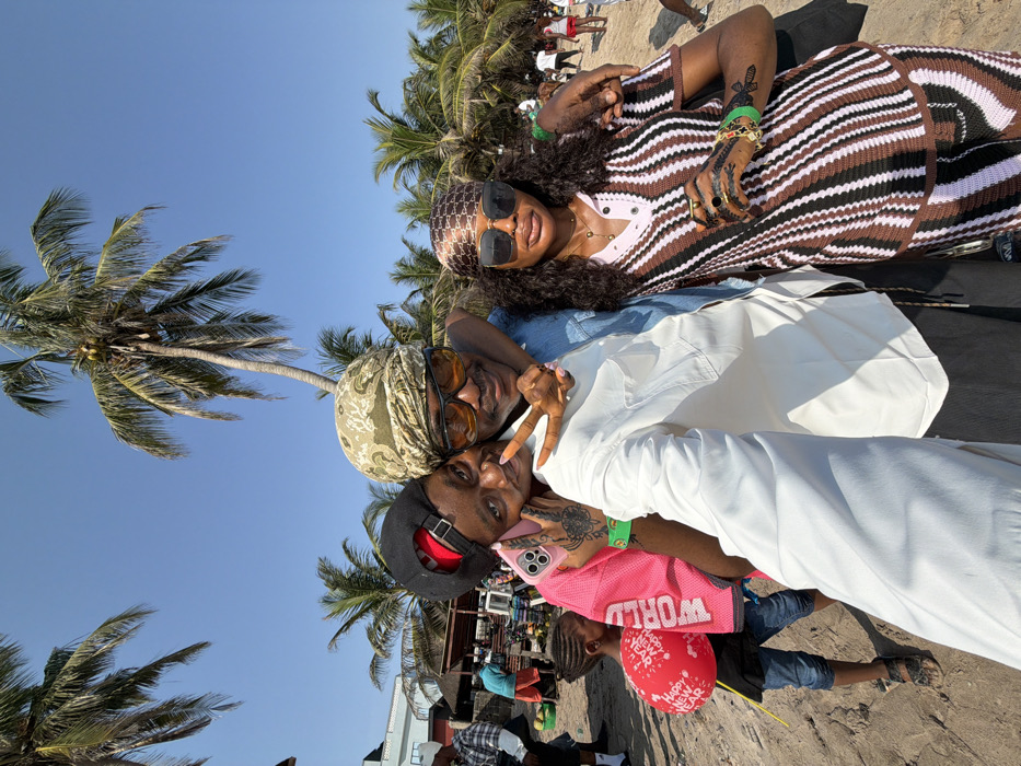 This image captures three adults posing for a selfie on a sandy beach under a clear blue sky, located in Mopo Onijebu, Nigeria.
In the foreground, a man in the center wears a white long-sleeve shirt under a denim overshirt, a khaki-patterned headwrap, and orange-tinted sunglasses. He embraces the two individuals beside him. To his left, a person with a black baseball cap and a pink mesh top smiles, holding a pink smartphone case and displaying a peace sign with their right hand, which has orange-painted fingernails and visible tattoos on the wrist. A green wristband is on their left wrist. To the man's right, a woman with long dark curly hair wears a brown patterned headscarf, large dark sunglasses, and a brown, black, and light pink striped knit two-piece outfit. She has a gold necklace, multiple rings, and a dark, intricate tattoo resembling henna on her right hand, along with a green wristband.
The background reveals a bustling beach environment with numerous tall palm trees, some bearing coconuts. Other individuals are scattered across the sandy terrain. To the far left, a young child in a dark shirt and blue jeans holds a deflated red balloon bearing the text "HAPPY NEW YEAR WORLD." Beyond, market-style stalls or vendors are visible, displaying items such as green coconuts, and a light-colored building with a blue roof stands further back.