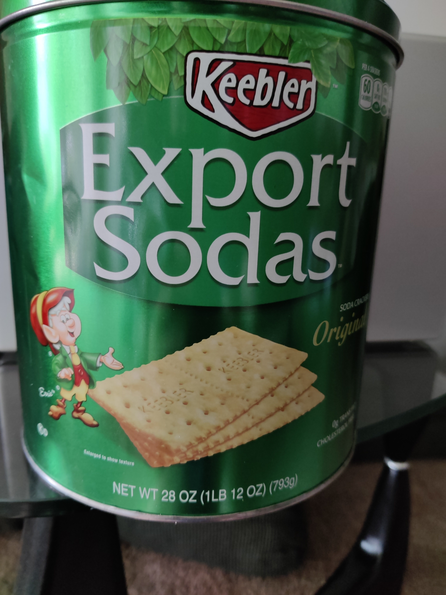 A close-up, eye-level shot shows a green tin of Keebler Export Sodas. The tin is mostly green with white lettering and a picture of a Keebler elf on the left side. The word "Keebler" is in a red and white oval logo at the top center. Below that, "Export Sodas" is written in large white letters. Three stacked crackers are visible in the lower right corner of the tin. The tin sits on a reflective glass surface, and a dark, curved object is visible in the lower right. The image appears to be taken indoors, and there is no indication of a specific location in Lower Paxton Township, United States of America.