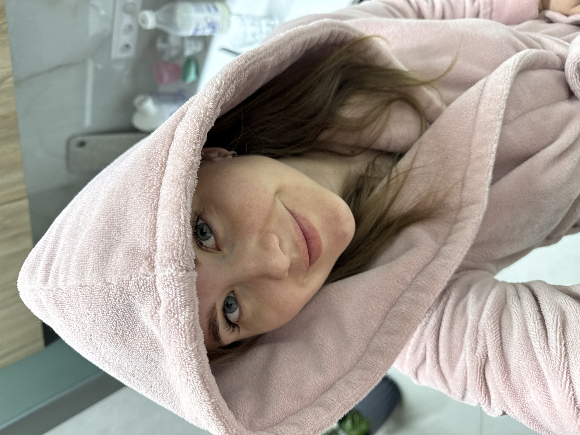 A woman with fair skin and blue eyes is seen from the chest up. She is wearing a light pink hooded robe. Her head is turned to the left, and she is looking directly at the camera with a slight smile. Light brown hair is visible around her face. The background includes a mirrored wall with various bottles and containers visible, and a small potted plant. The image orientation is rotated slightly clockwise. It is not possible to ascertain the location is Nestiukovo, Russia from this image.